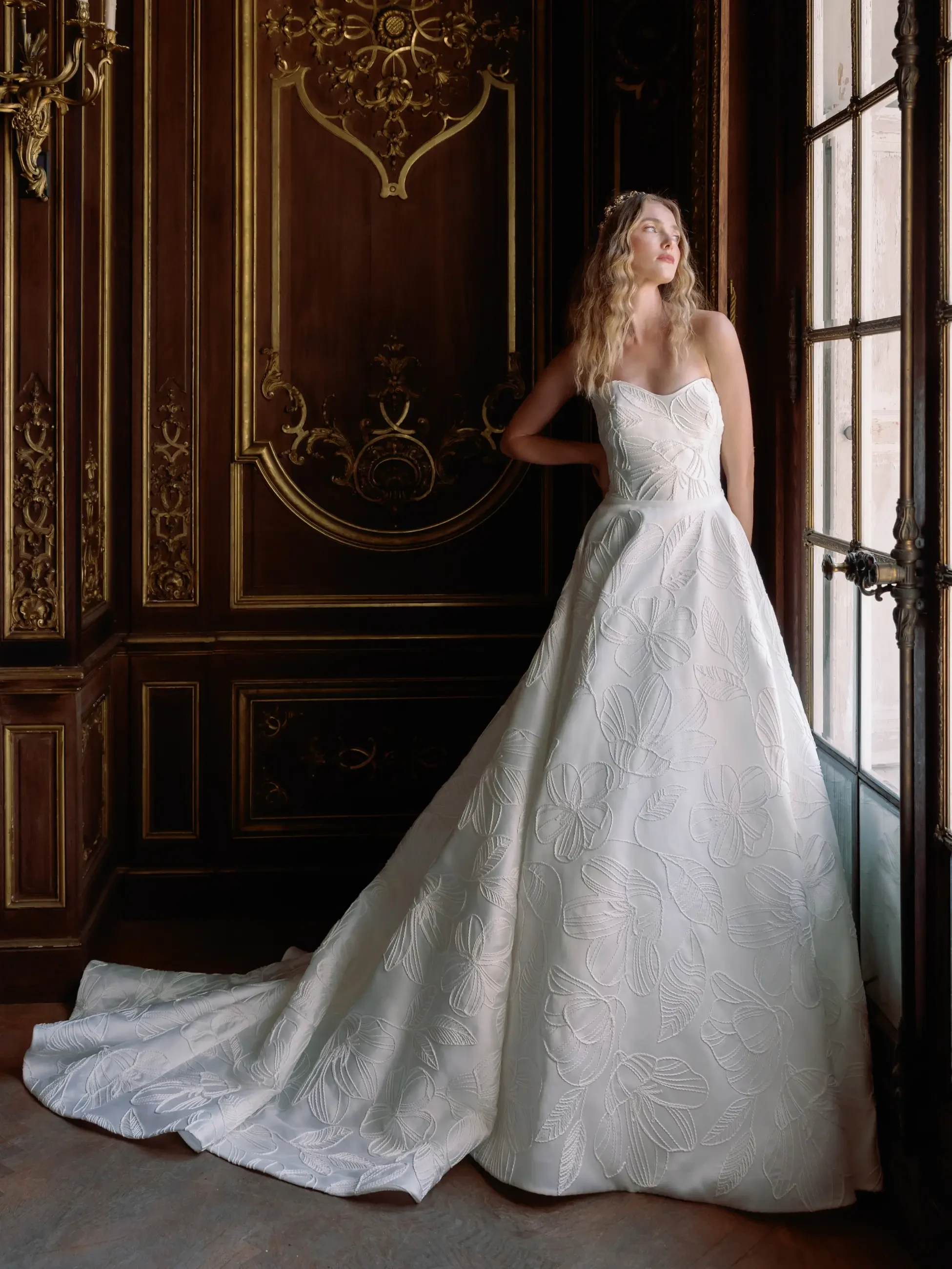 A woman in a strapless white gown with floral patterns stands by an ornate, dark wood wall with gold details, gazing thoughtfully out a window.