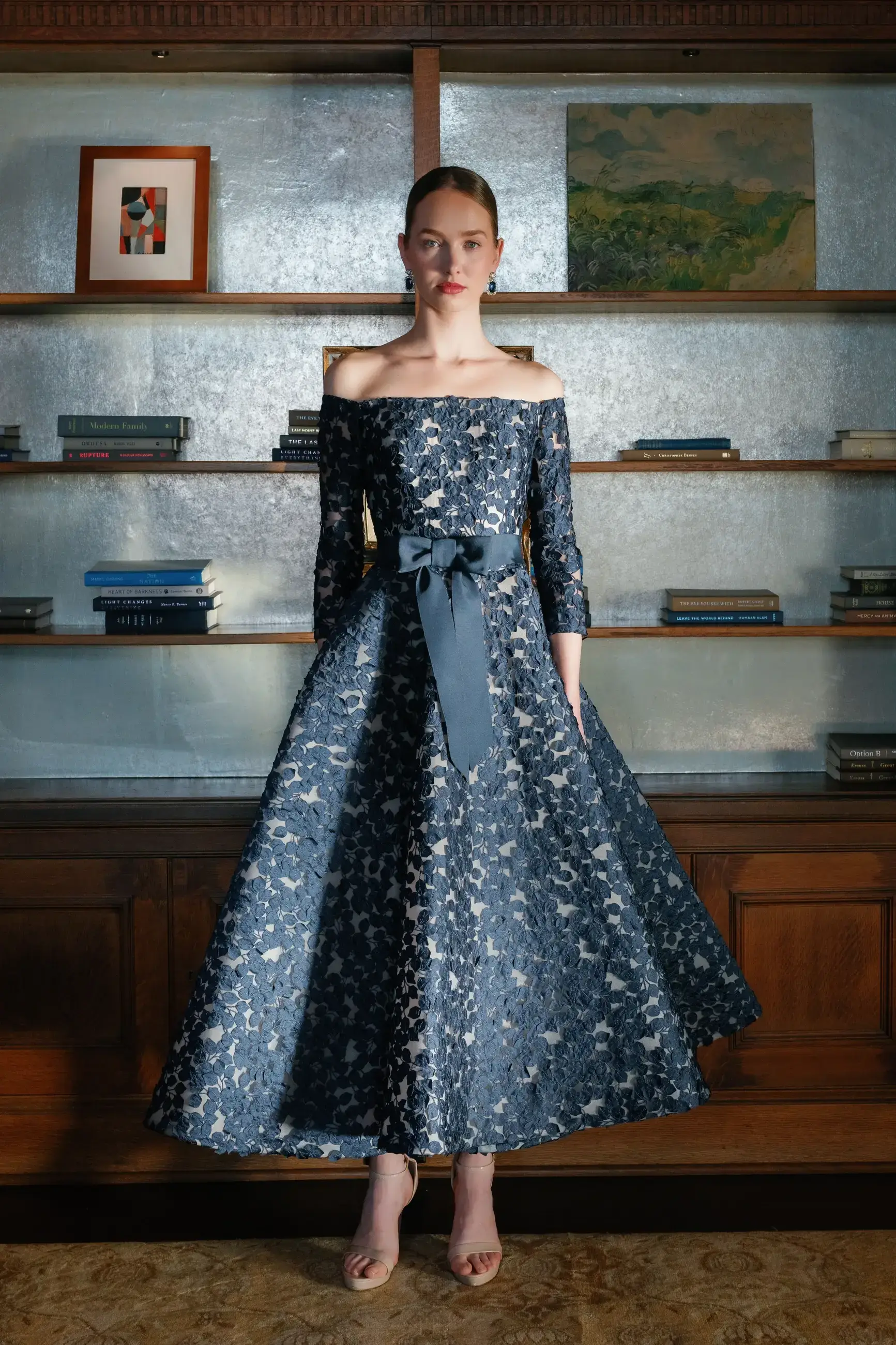 A model wearing an off-the-shoulder, navy blue dress with a bow at the waist, standing gracefully in front of a bookshelf filled with books and framed artwork.