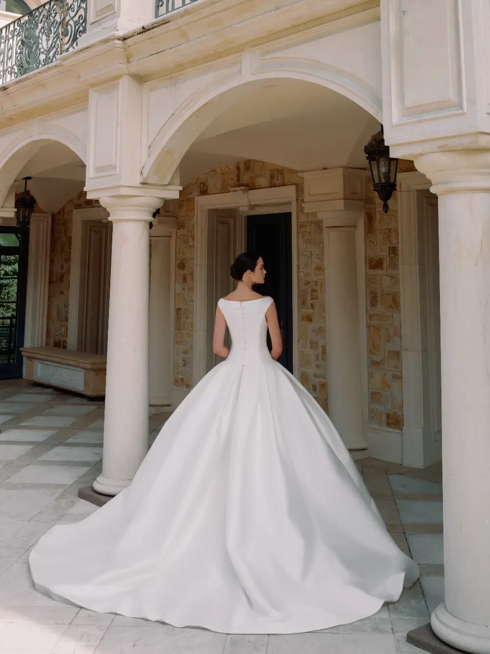 A bride wearing a simple, elegant white wedding gown with a full skirt and a netted veil, standing in an architectural setting with columns.
