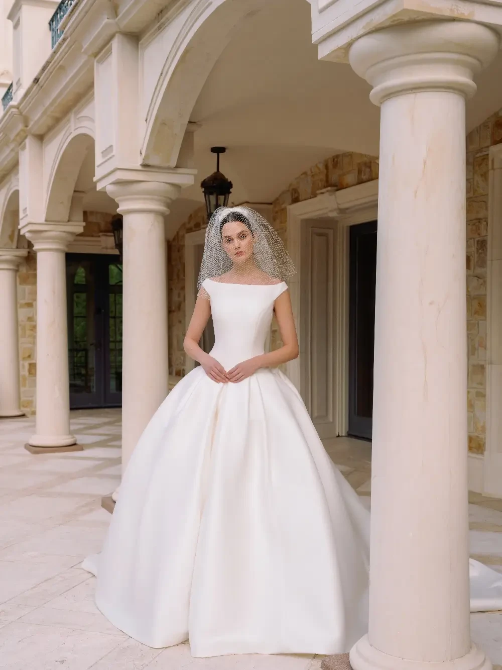 A bride wearing a simple, elegant white wedding gown with a full skirt and a netted veil, standing in an architectural setting with columns.