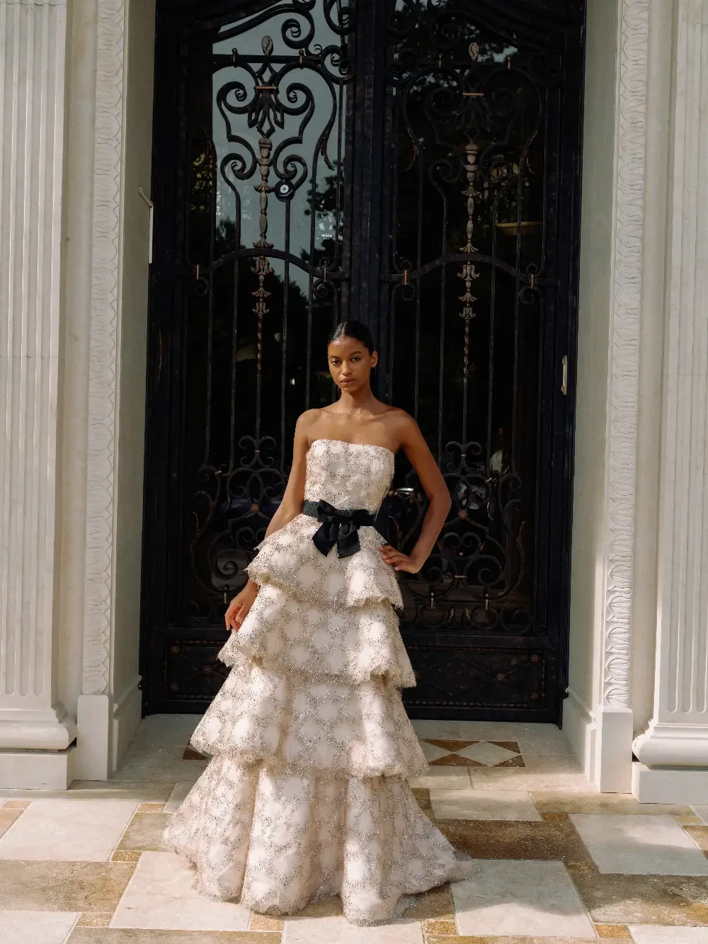 A model wearing a strapless, tiered white gown with ornate detailing and a black bow, standing in front of an ornate door.