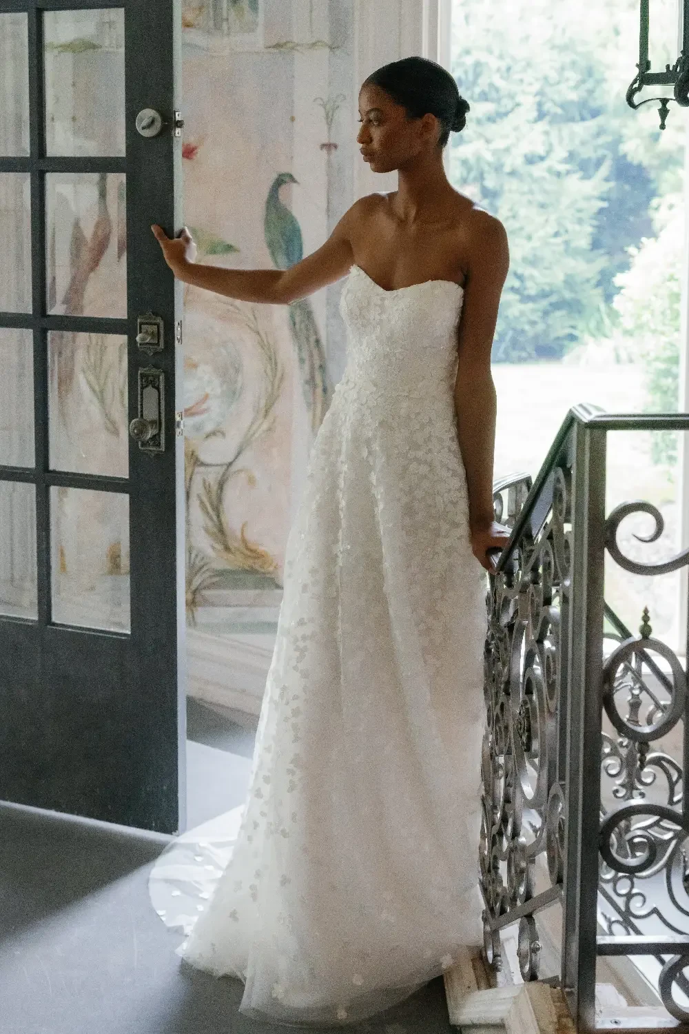 A model stands elegantly in a strapless white wedding dress with floral embellishments, posing in a sunlit room with large windows.