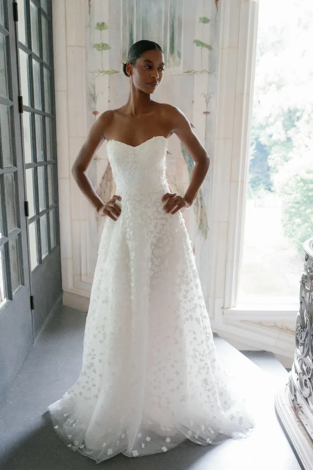 A model stands elegantly in a strapless white wedding dress with floral embellishments, posing in a sunlit room with large windows.