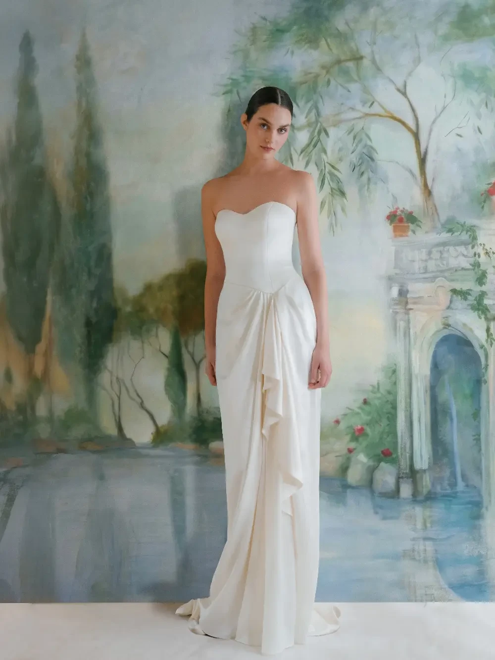 A model wearing a strapless white gown stands in front of a painted background depicting a serene landscape with greenery and an archway.