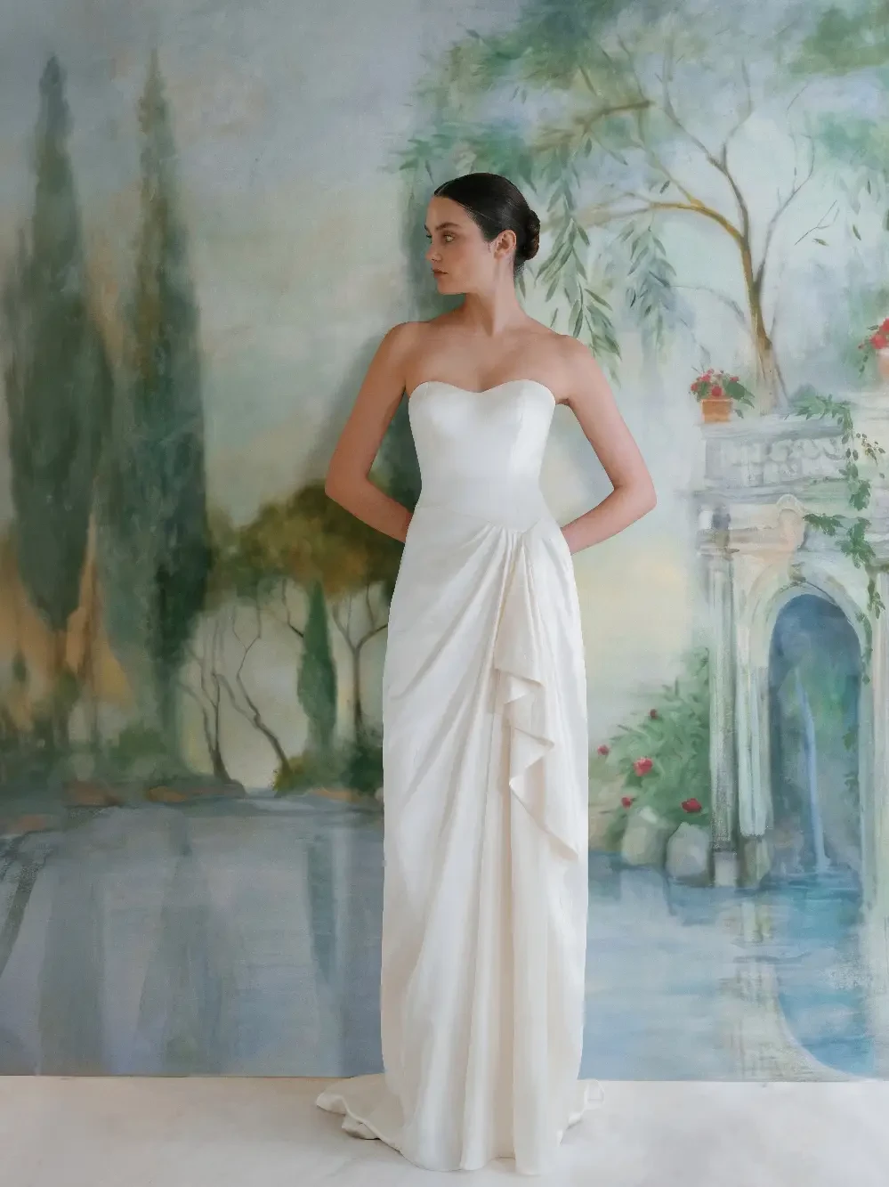 A model wearing a strapless white gown stands in front of a painted background depicting a serene landscape with greenery and an archway.
