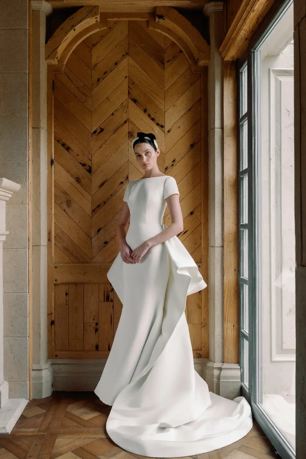 A woman wearing a white dress with a unique textured pattern stands against a wooden wall.