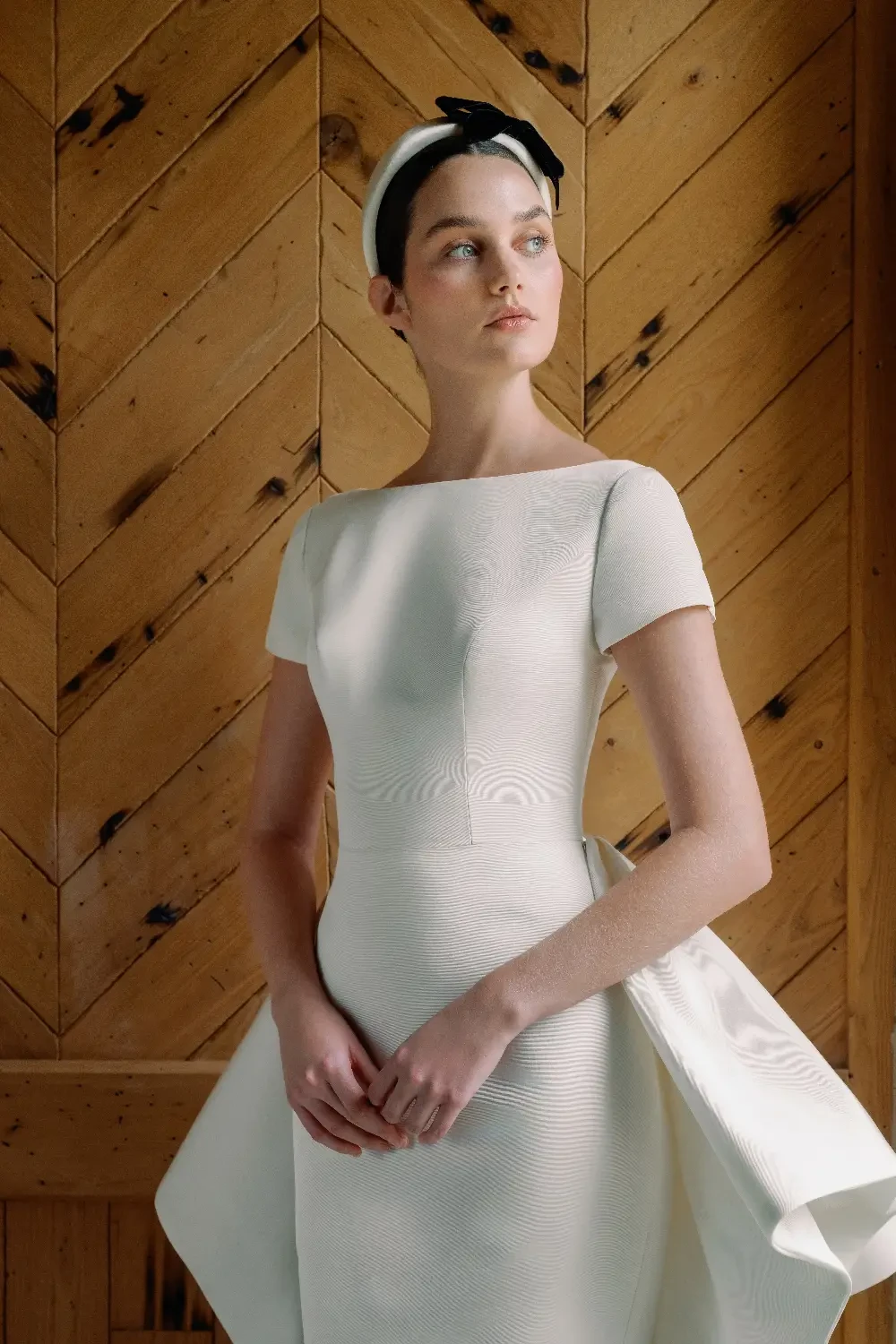 A woman wearing a white dress with a unique textured pattern stands against a wooden wall.
