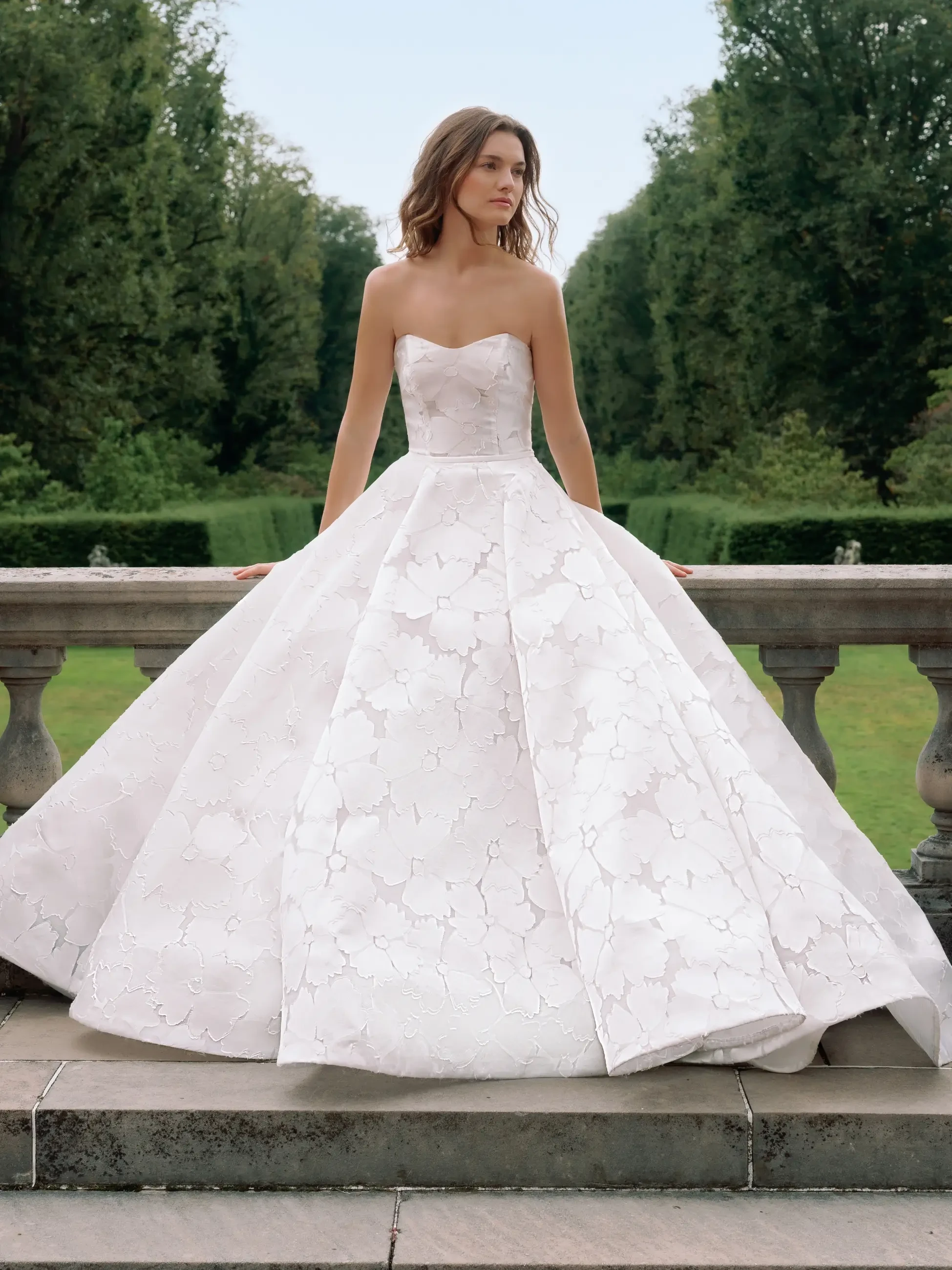 Elegant woman in a strapless, floral-patterned white gown stands on a terrace. She gazes pensively to the side, flanked by lush green gardens.