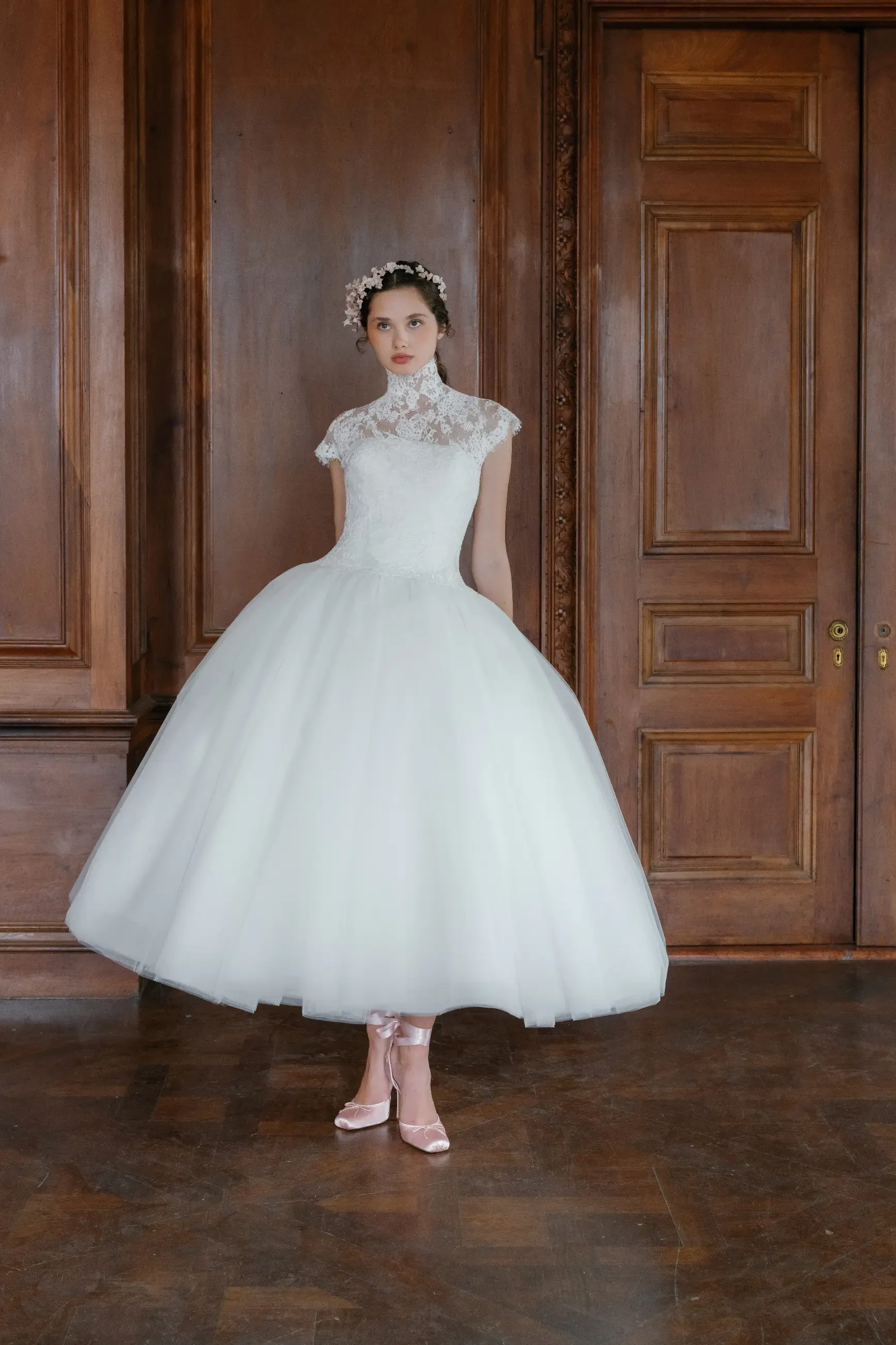 A model wearing a white, knee-length ball gown with lace details and a high neckline, standing on a wooden floor in an elegant room with wooden paneling.