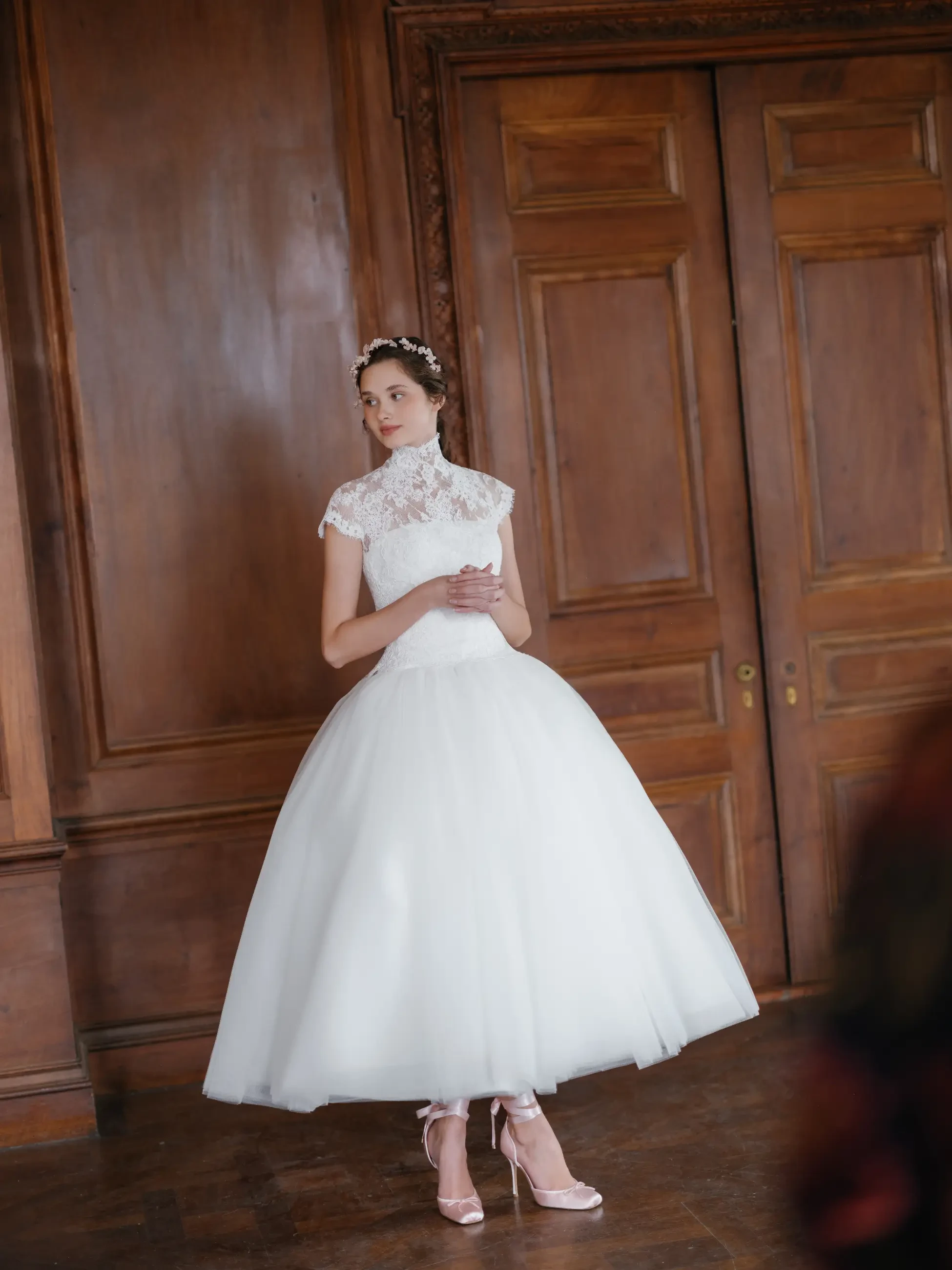 A woman in a white dress with a fitted bodice and full skirt stands in front of wooden paneling. She has her hair styled and is wearing a floral headpiece. The setting appears to be an elegant indoor space.