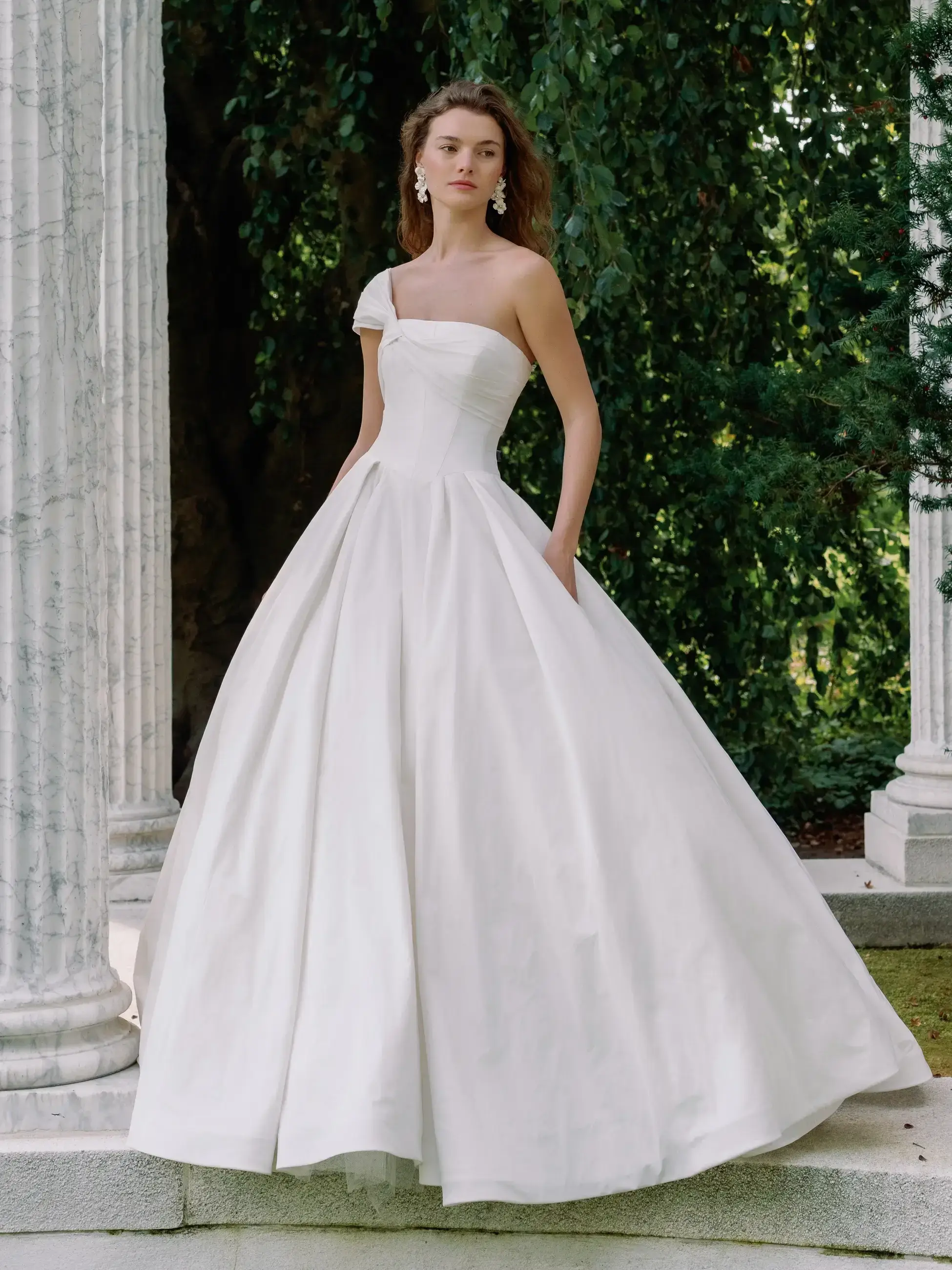 A woman wearing a strapless white ball gown with one shoulder strap, standing gracefully in a garden setting near classical columns.