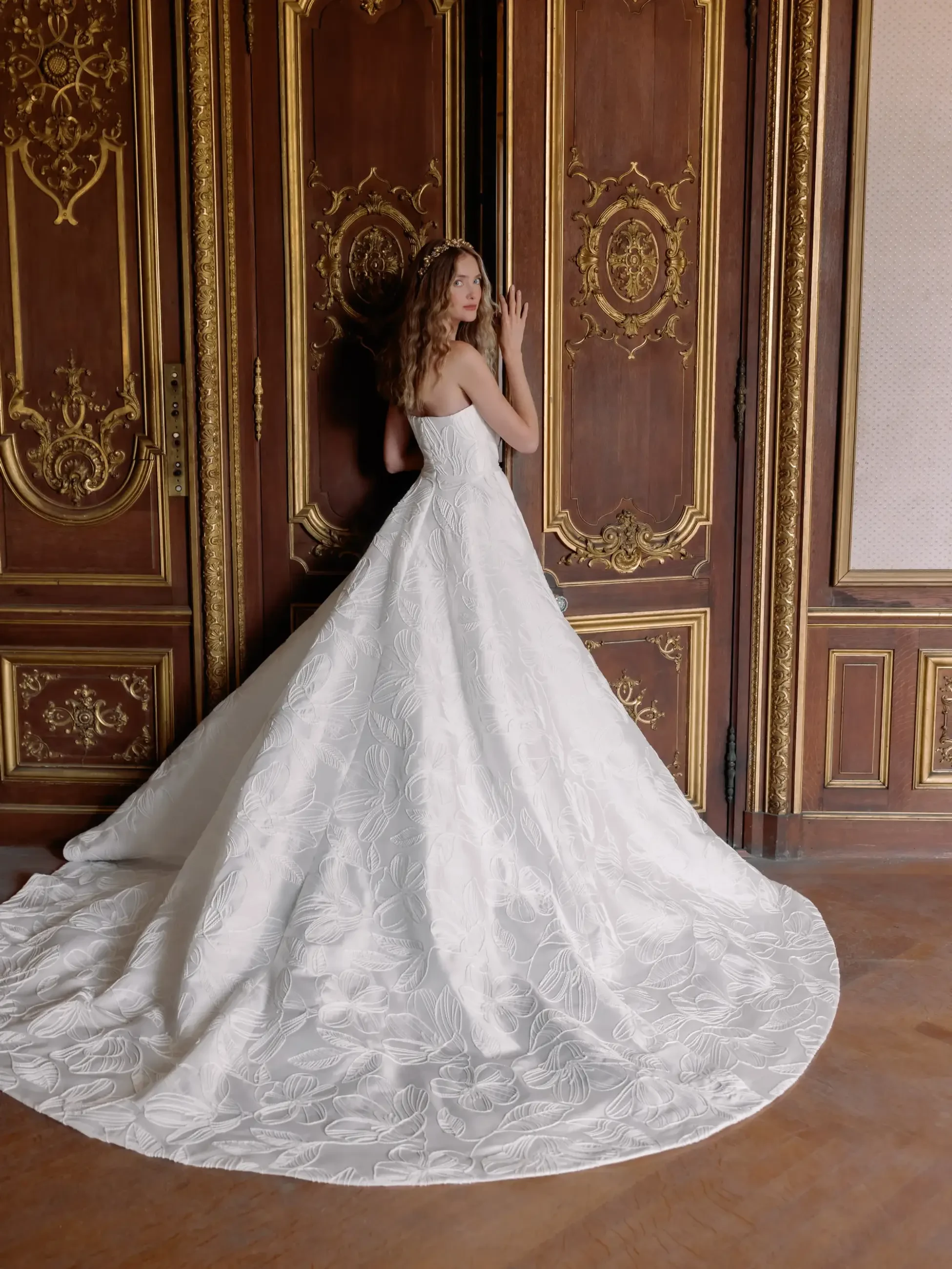 A woman in an elegant white bridal gown stands by ornate, dark wood doors with gold detailing. The scene evokes a sense of classic romance.