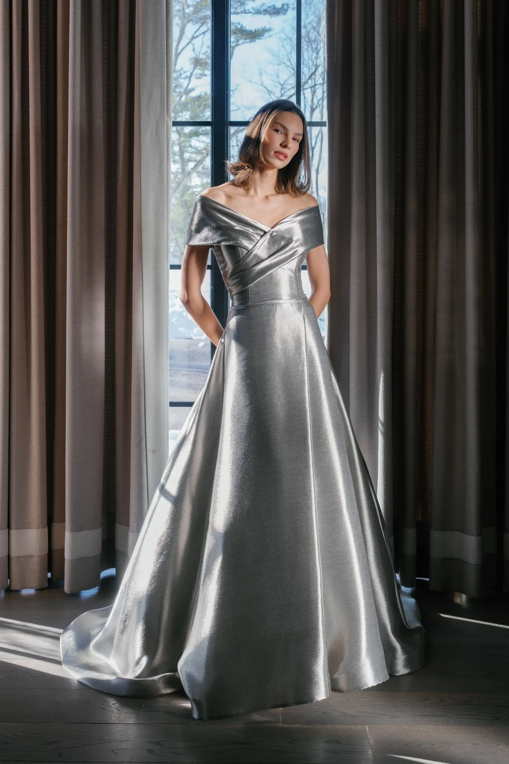 A woman in a silver off-shoulder gown stands in front of large windows, with soft natural light illuminating the scene. The gown has a fitted bodice and a flowing skirt.