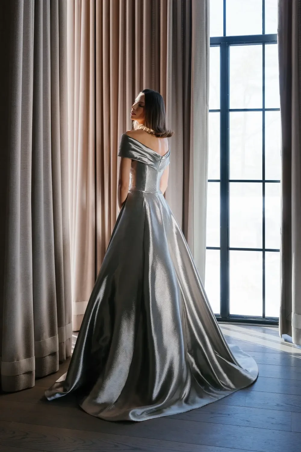 A woman wearing a silver gown, standing with her back to the camera in front of large windows with light curtains.