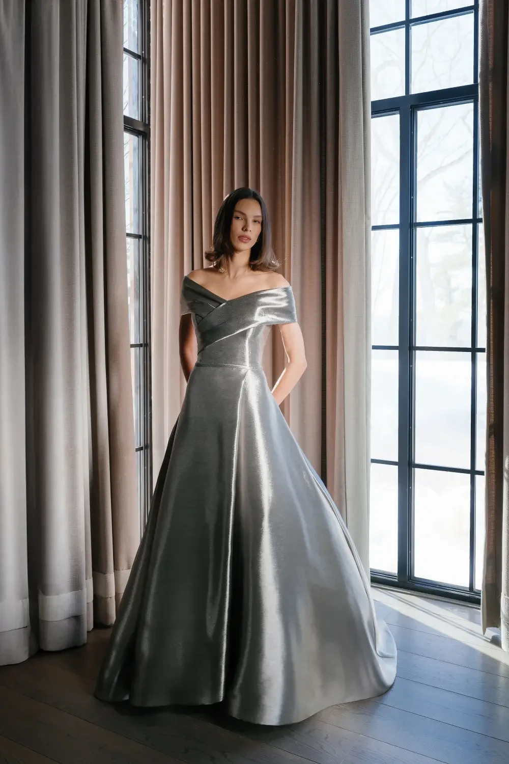 A person wearing a silver off-the-shoulder gown stands near a window with sheer curtains.