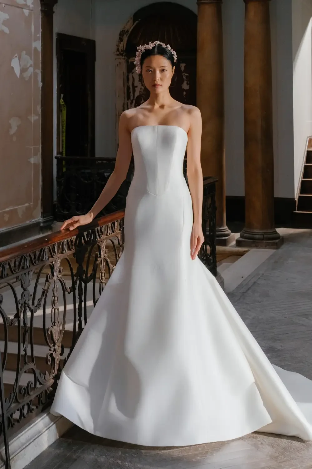 A model wearing a strapless, fitted white wedding gown stands gracefully in an elegant interior.