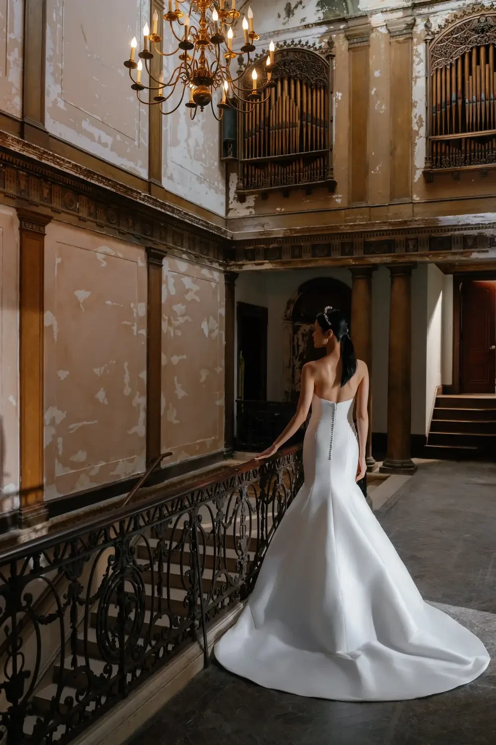 A woman in a white wedding dress stands on a staircase, viewed from behind, in an ornate but aged interior with chandeliers and peeling walls.