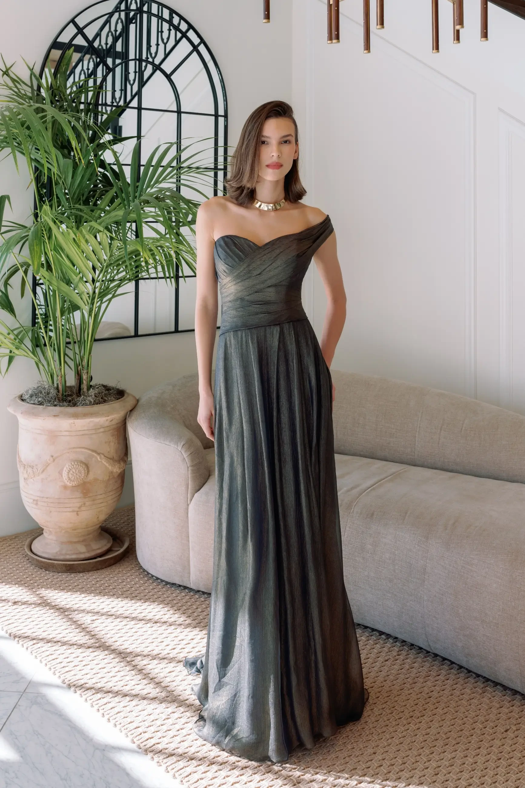 A woman wearing an elegant off-shoulder gown stands in a well-lit room with a large window and a potted plant.