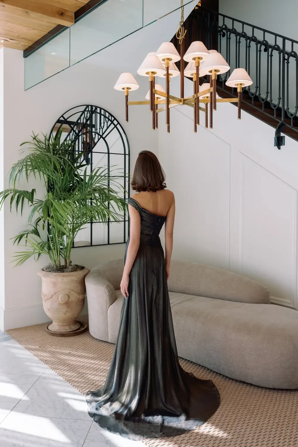A woman in a black dress stands with her back to the camera, in a stylish interior featuring a staircase, a large plant, and modern light fixtures.