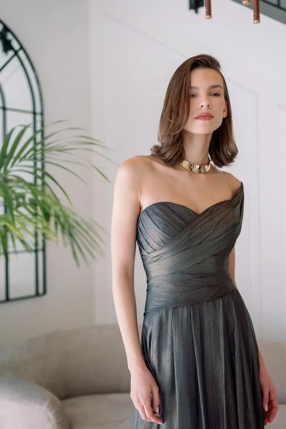 A woman in a one-shoulder, gray dress with a textured design, standing against a light-colored wall with a plant and a gray sofa in the background. She is wearing a gold necklace and has shoulder-length hair.