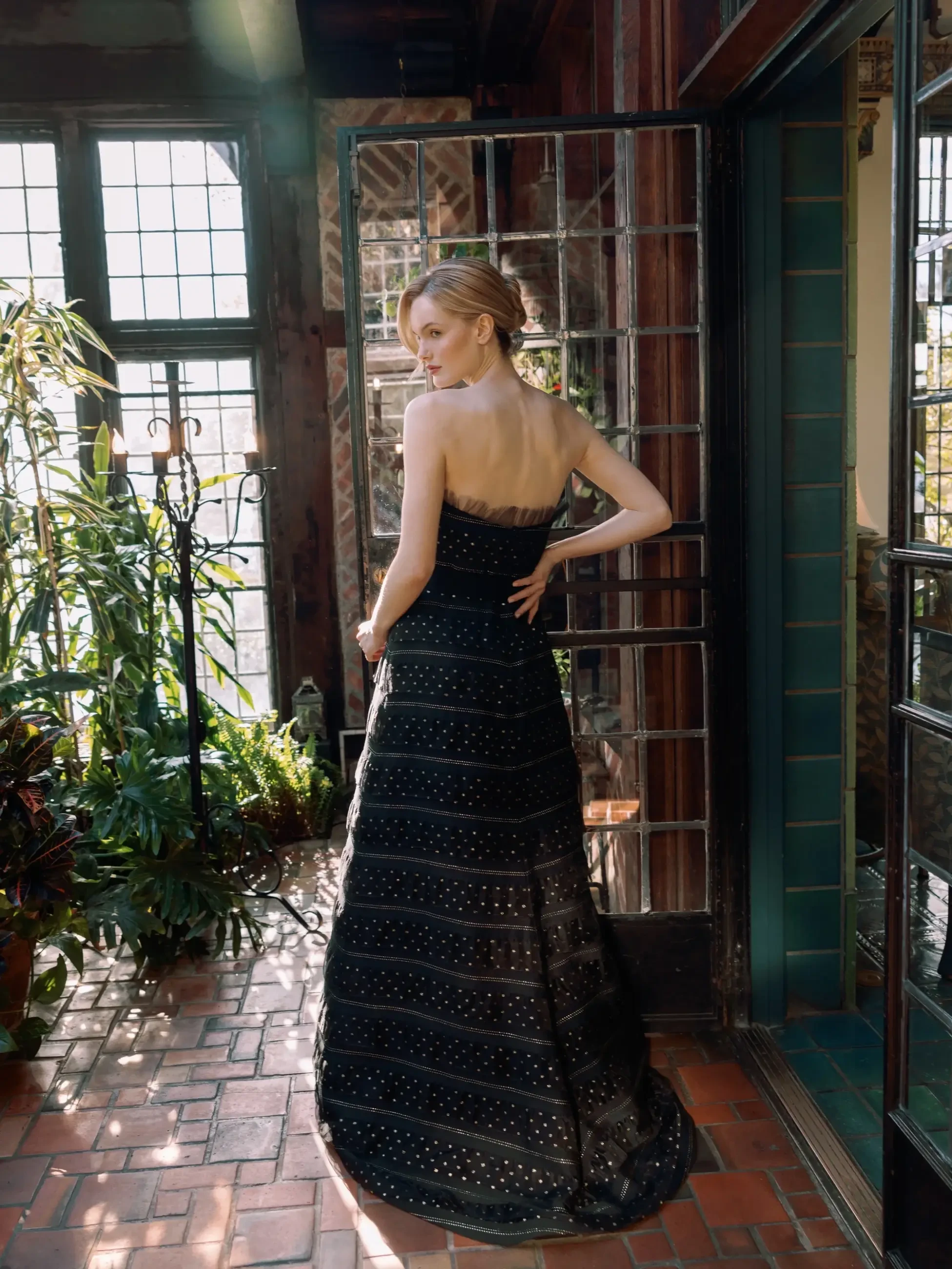 A woman in an elegant, strapless black gown stands on a sunlit brick patio. She is near large windows, surrounded by lush green plants.