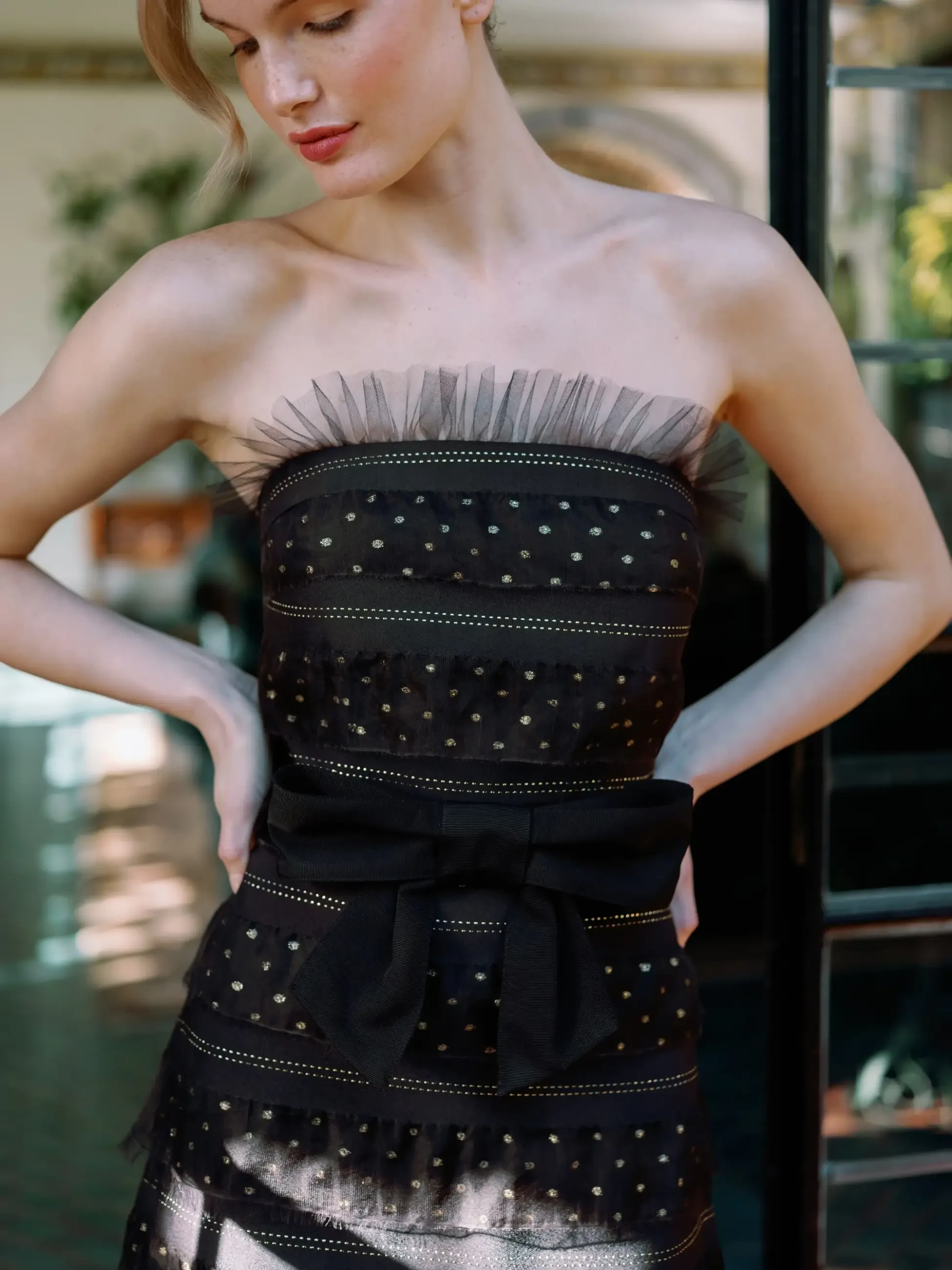 A woman in a strapless black dress with delicate polka dot and tulle details stands confidently. Her hands rest on her hips, and soft light highlights her elegant pose.