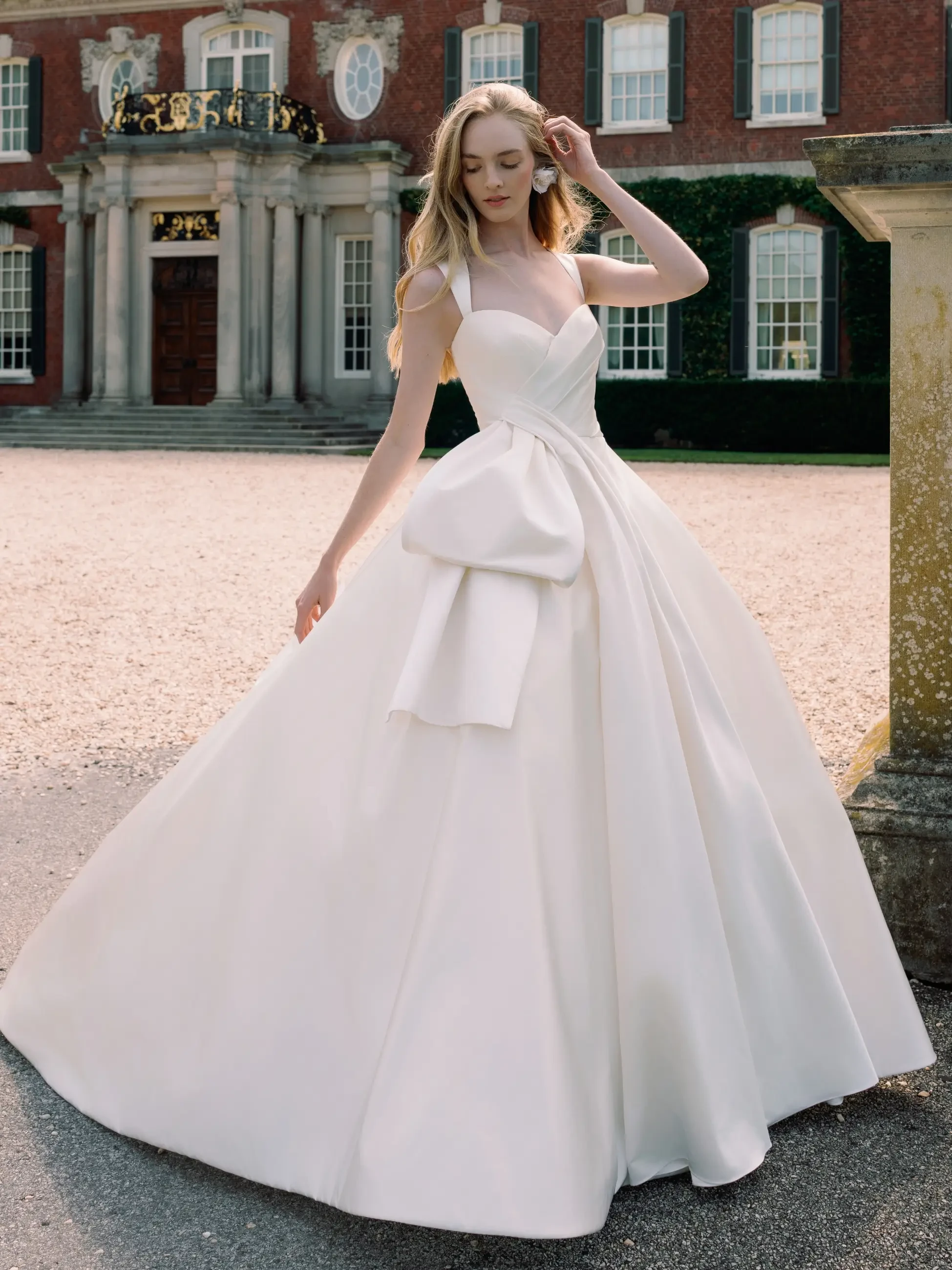 A woman in an elegant white wedding gown poses in front of a grand manor with columns. The setting is serene and sophisticated, conveying romance.