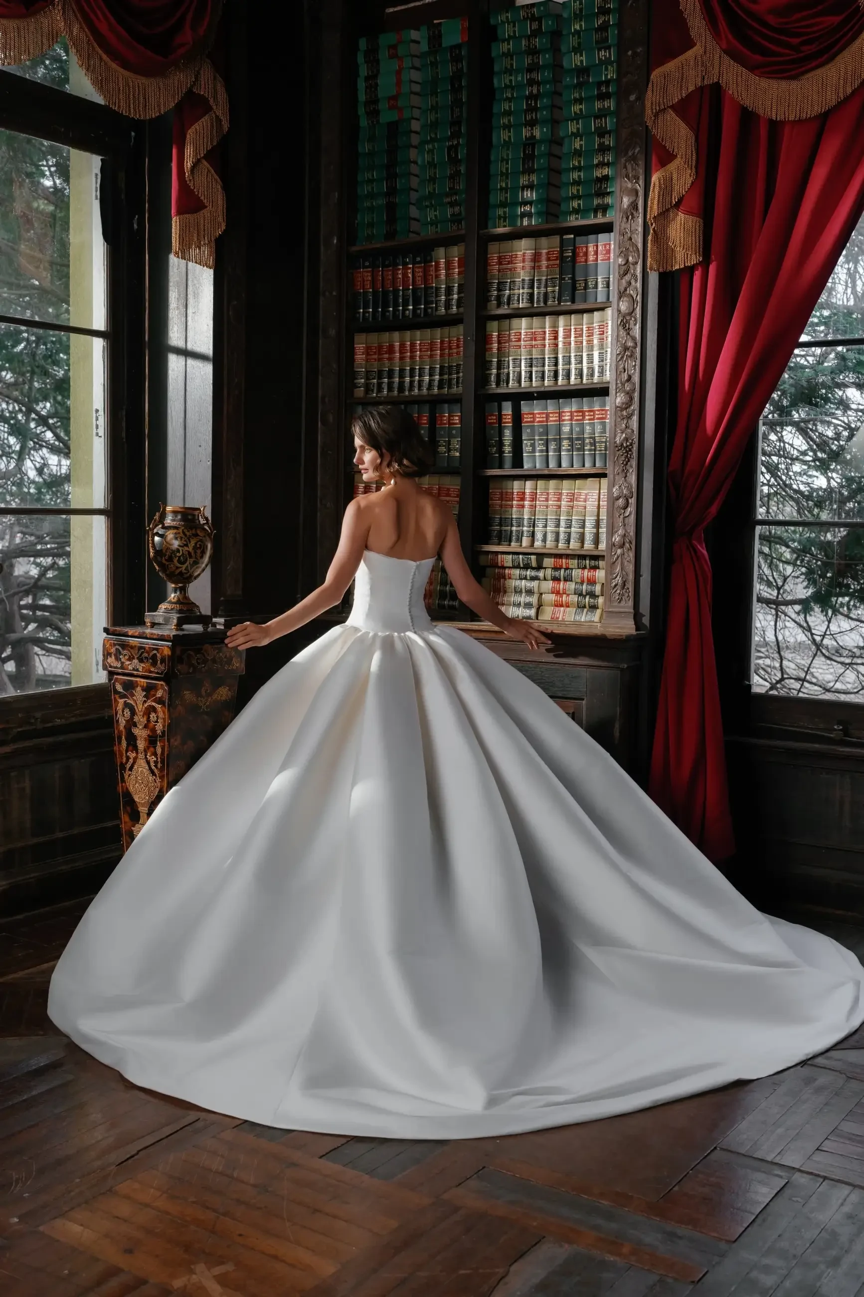 A woman in a white ball gown stands with her back to the camera, showcasing the gown's full skirt, in a richly decorated room with bookshelves and red curtains.