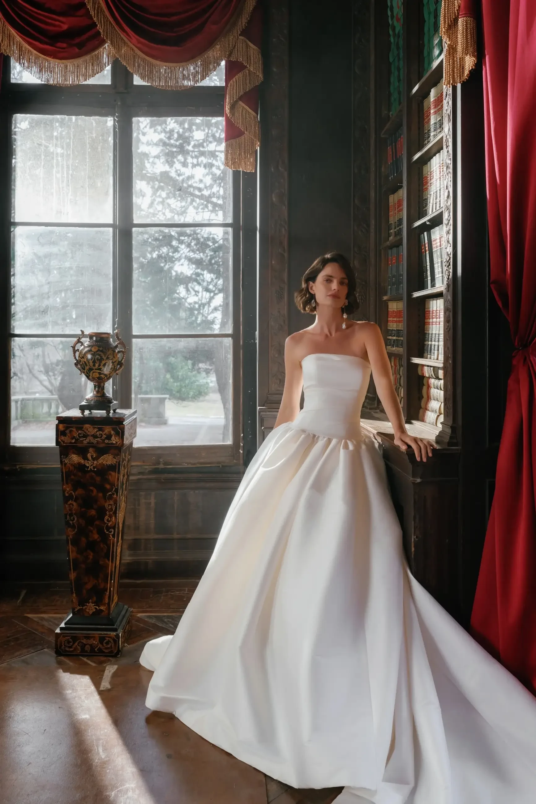 A woman in a strapless white wedding gown stands elegantly beside a large window, with bookshelves and red drapes in the background.