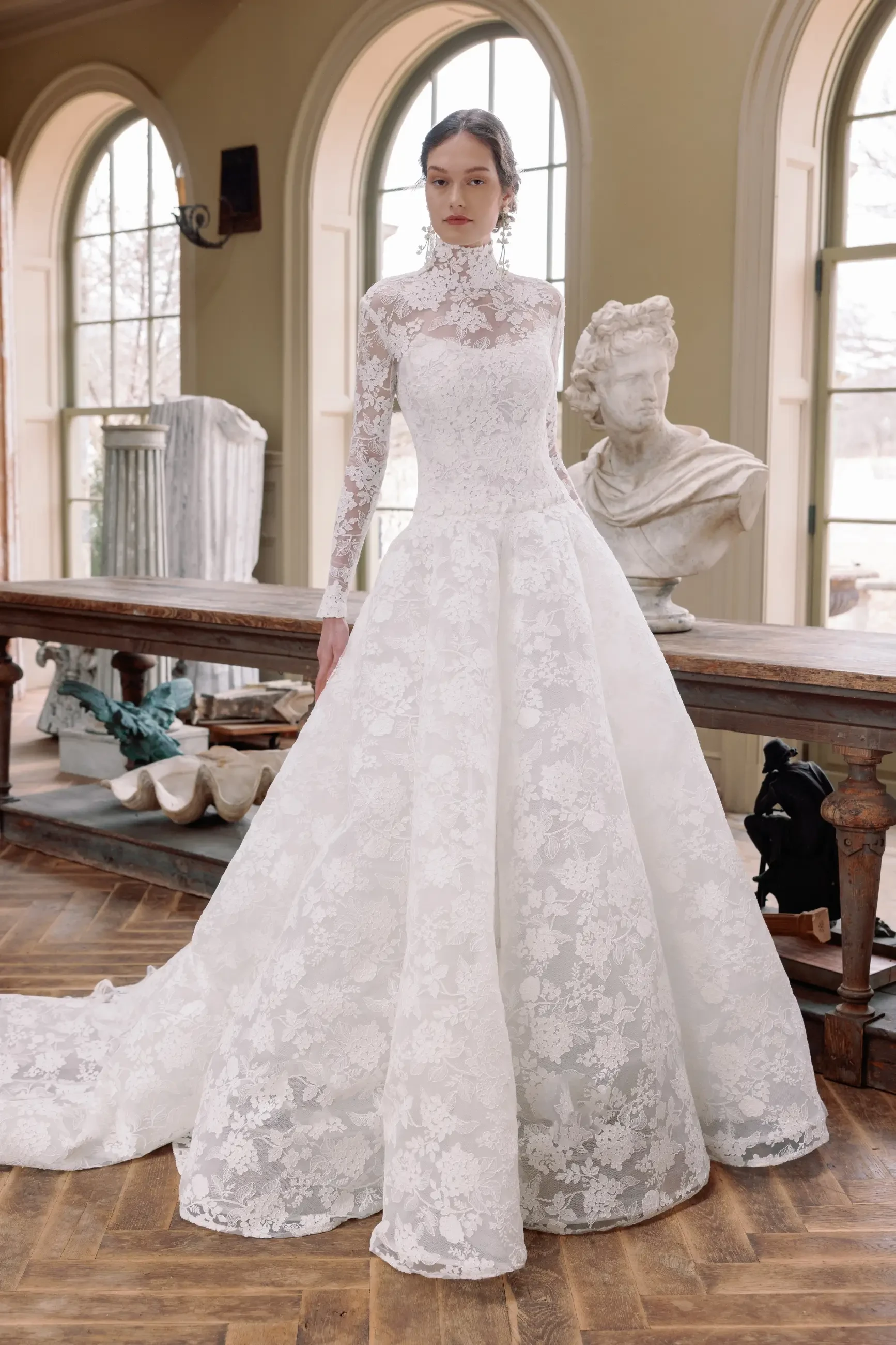 Model is wearing a ball gown crafted from embroidered hydrangea lace, featuring a cat-eye neckline, a flattering drop waist, and a dramatic coned silhouette with a chapel-length train.