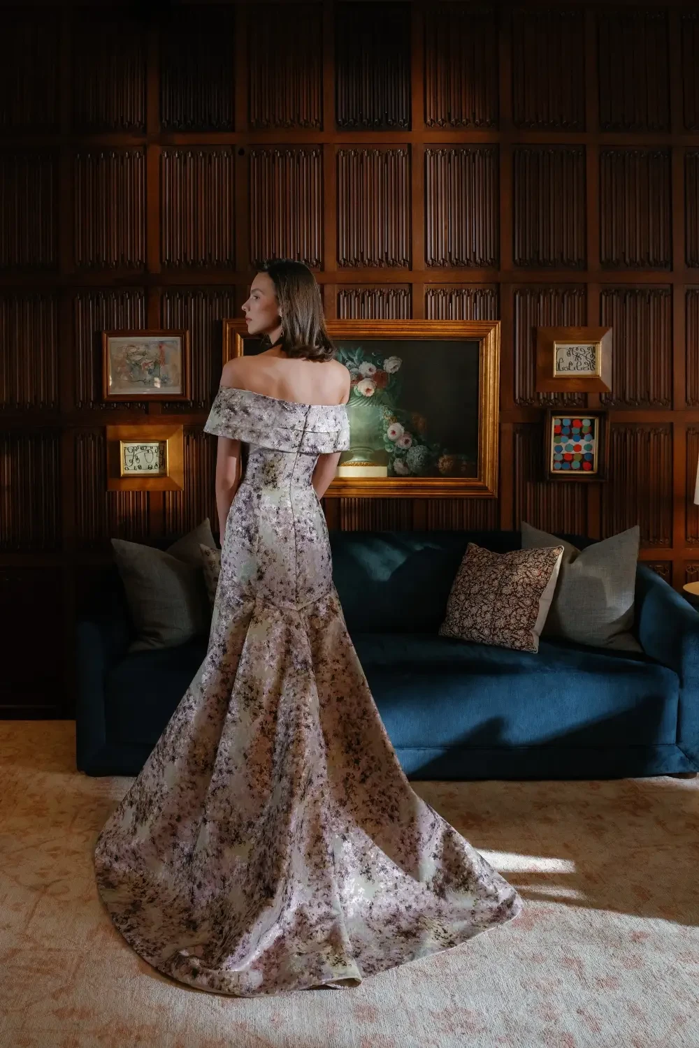 A model wearing an off-the-shoulder floral gown stands in a vintage room with wooden paneling, next to a dark blue couch and a decorative painting on the wall.