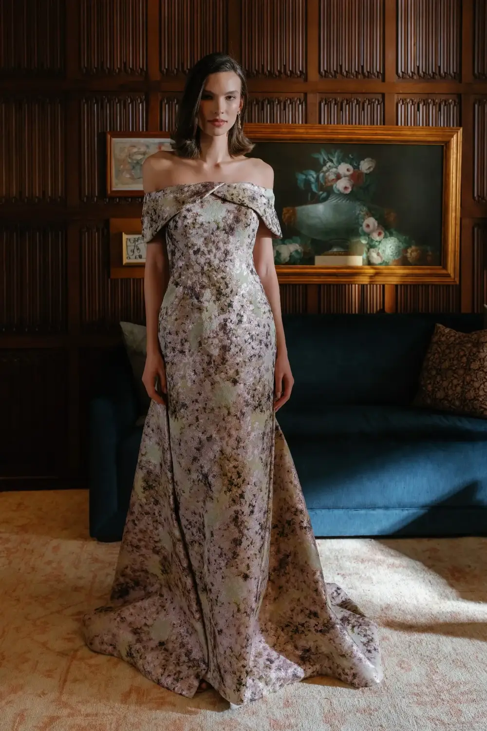 A woman in an elegant, off-the-shoulder floral gown stands in a room with wooden paneling, beside a dark blue sofa and a decorative painting on the wall.
