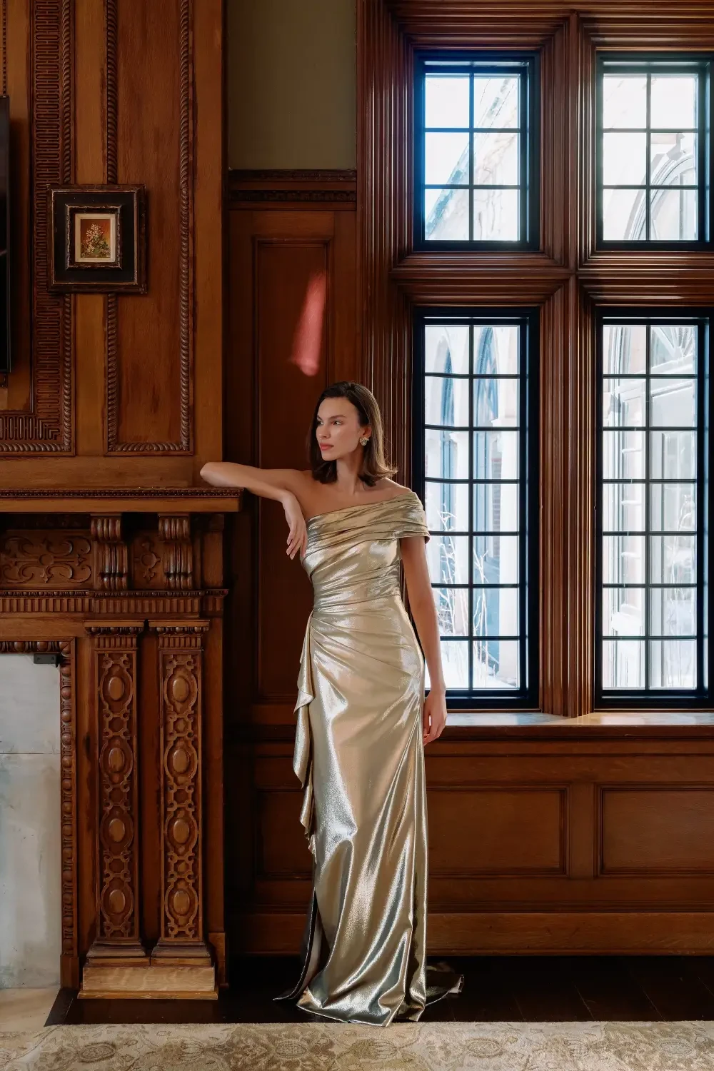 A woman in a gold dress stands gracefully beside an ornate wooden fireplace, with large windows and elegant wood paneling in the background.