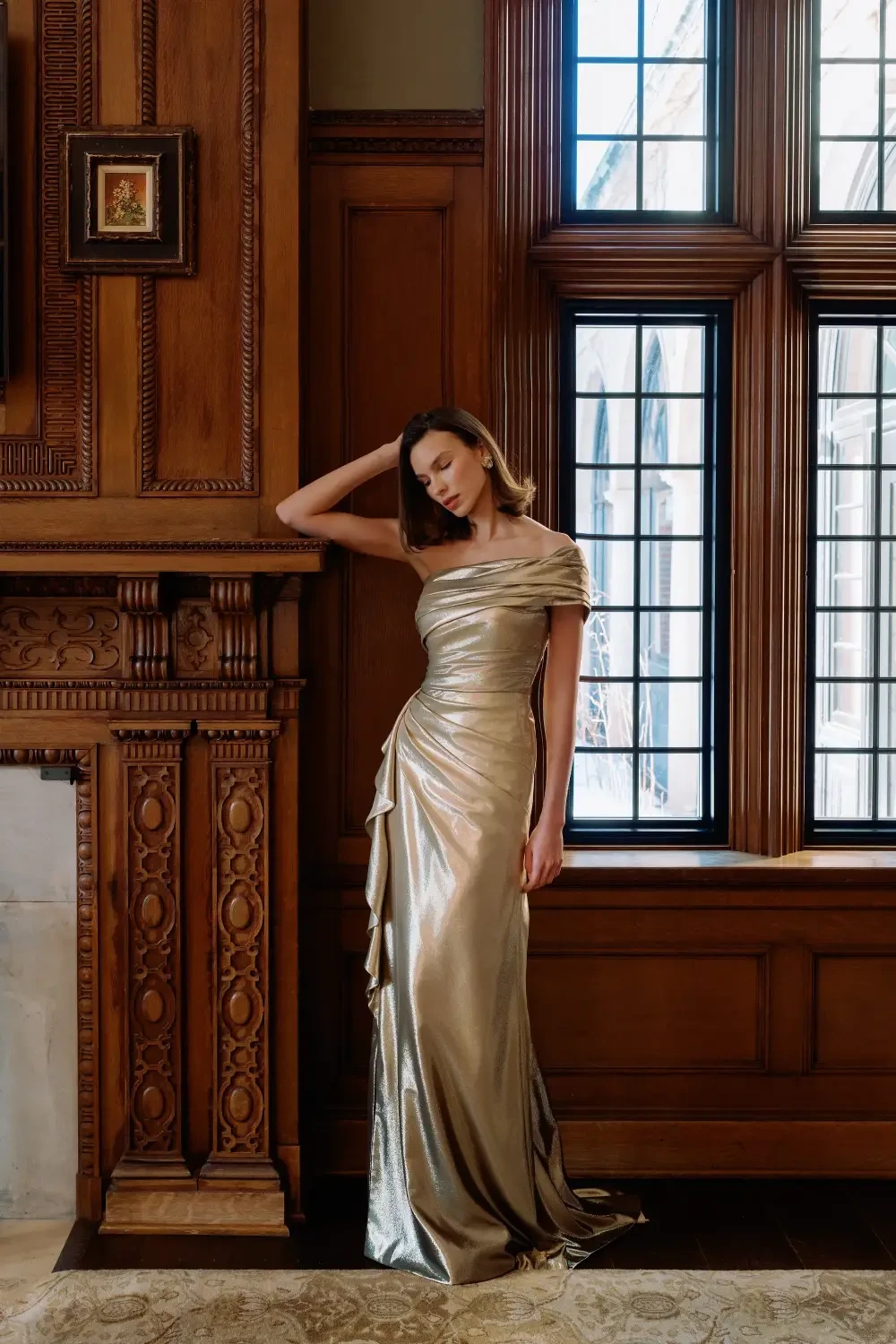 A woman in an elegant, gold gown leans against a wooden wall near large, paned windows, with a fireplace visible nearby.