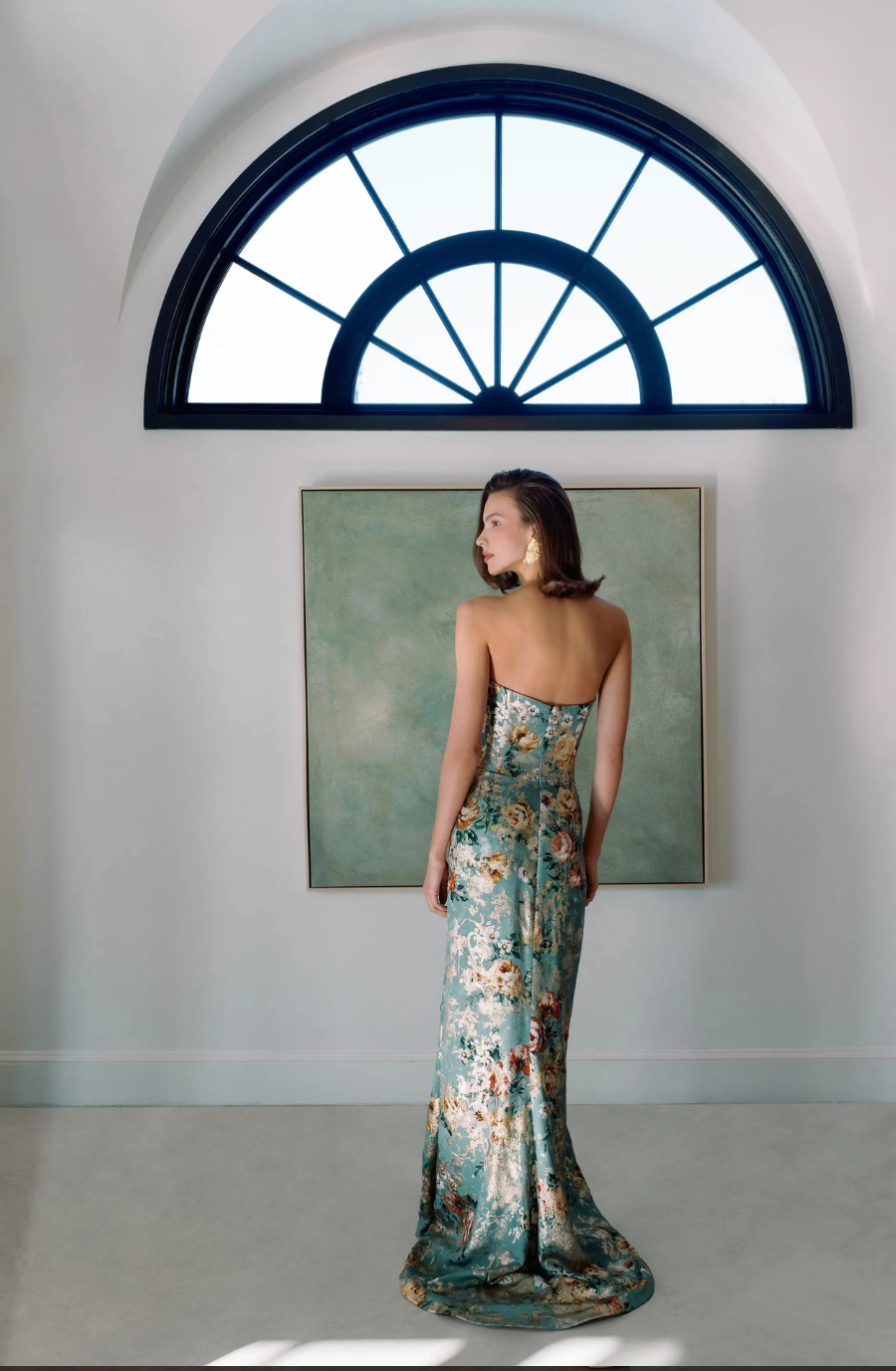 A woman wearing a floral gown stands with her back to the camera, facing a large window with an arch shape and a painting on the wall behind her.