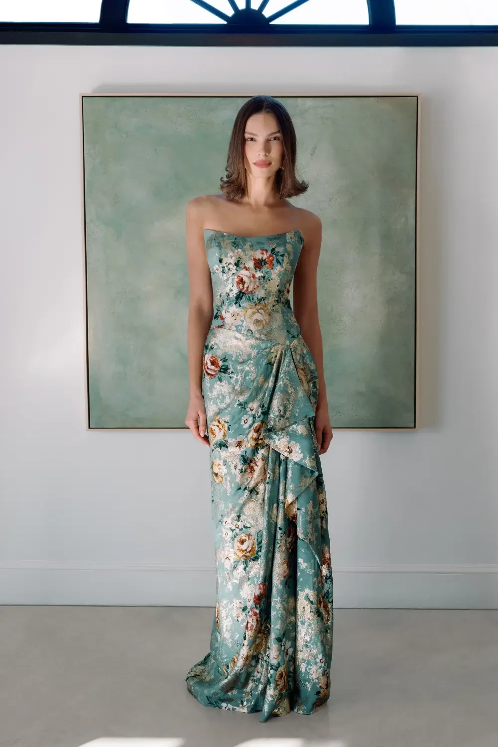 A woman is wearing a strapless floral dress, standing in front of a large painting. The dress is blue with intricate patterns, and the background includes a light-colored wall and a window.
