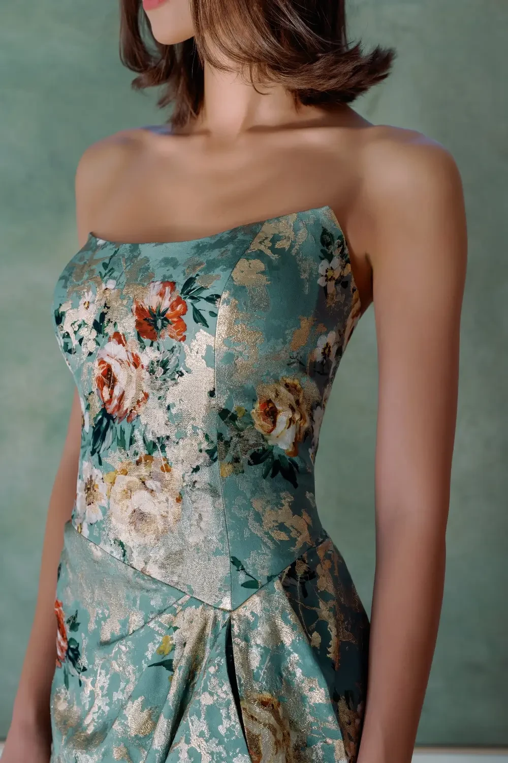 A woman stands in a floral-printed evening gown in front of a textured green painting.