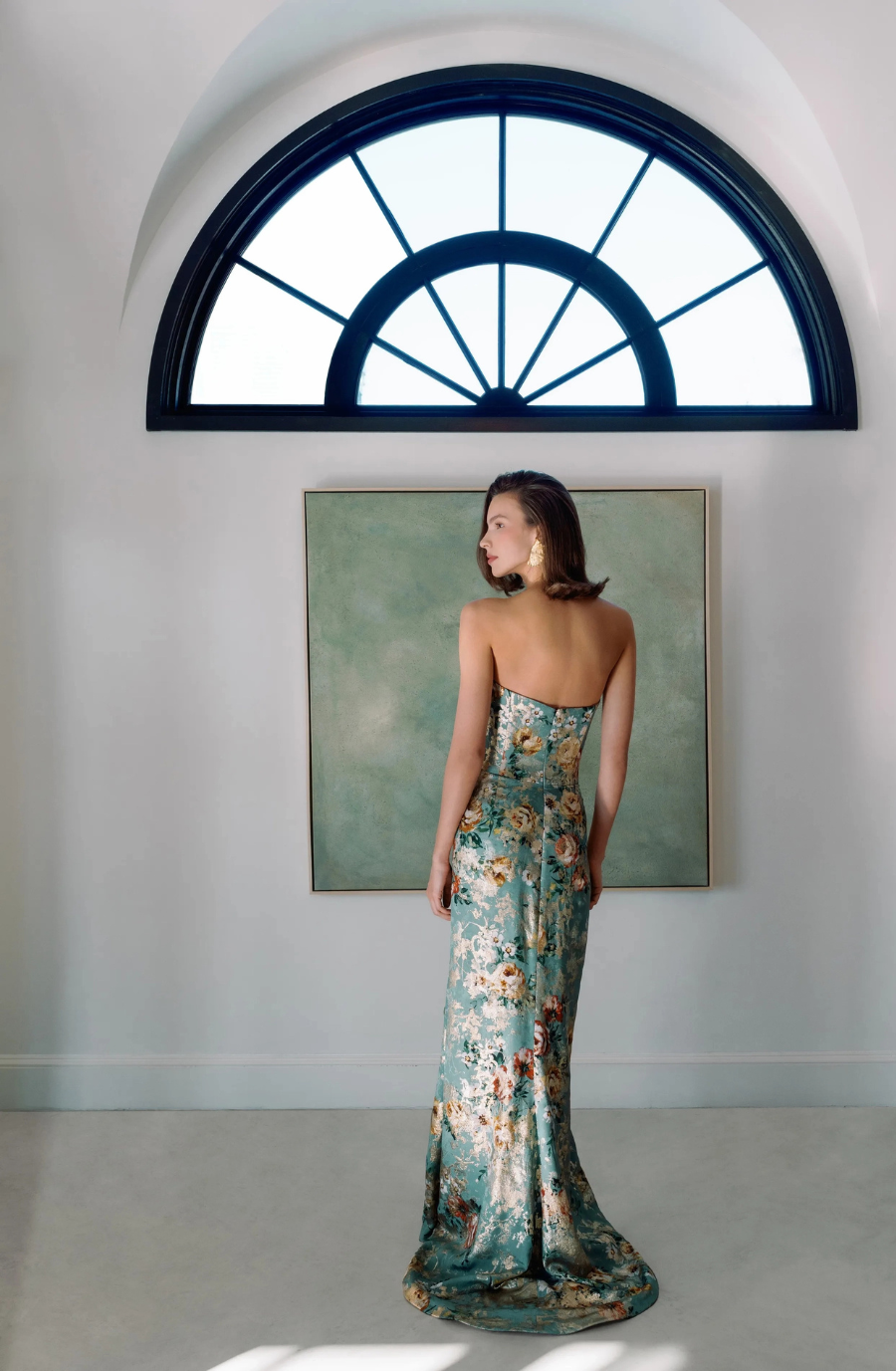 A woman wearing a floral evening gown stands with her back to the camera, facing a large arched window and a green artwork on the wall.