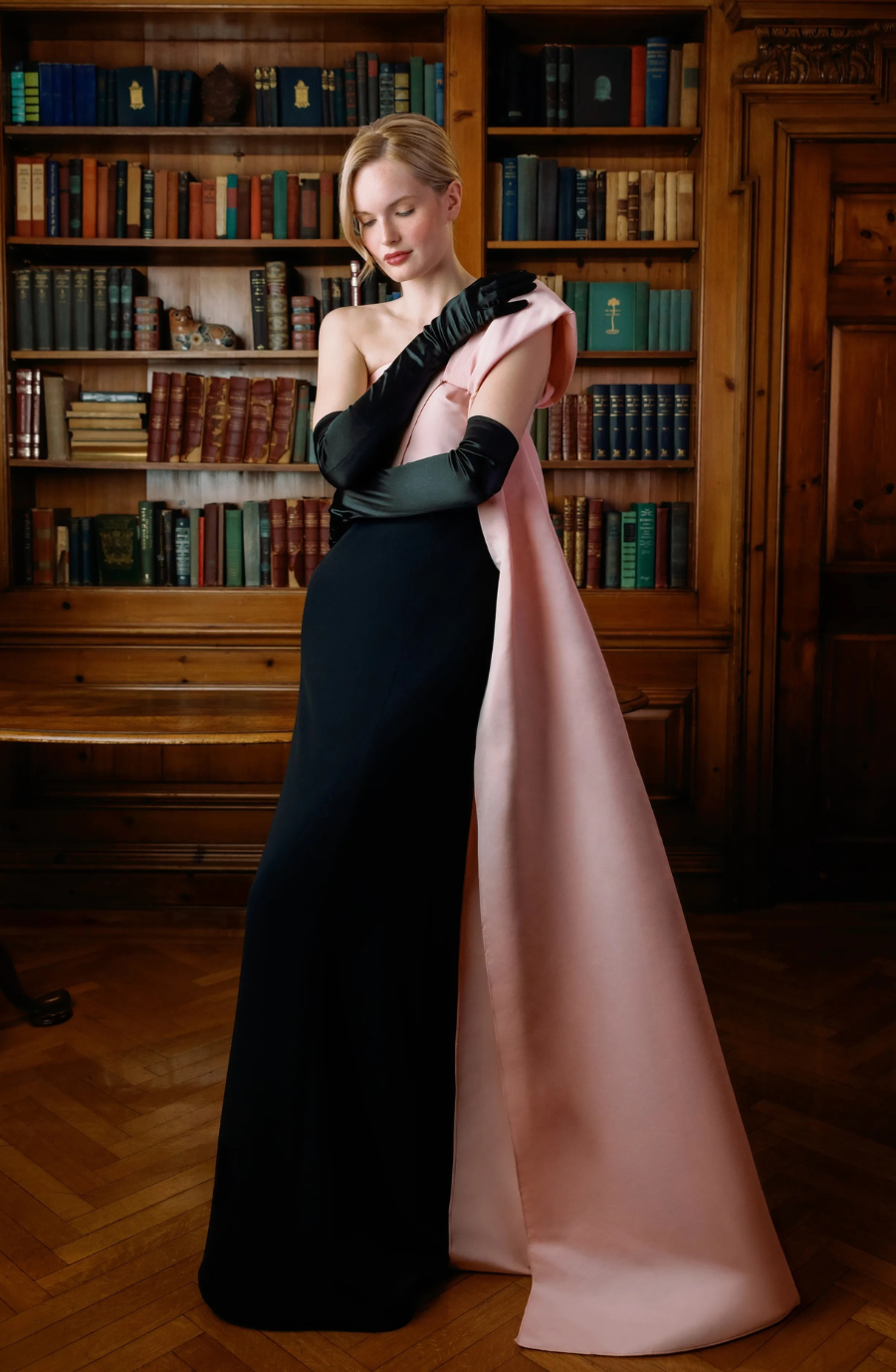 A woman in a sleek black gown with a pink satin train, wearing long black gloves, stands poised in a wooden library, exuding elegance and grace.