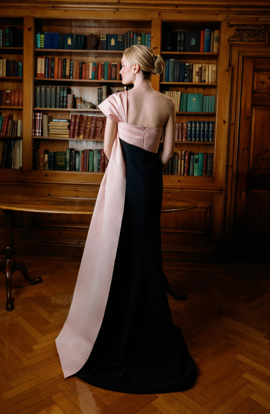 A woman in a sleek black gown with a pink satin train, wearing long black gloves, stands poised in a wooden library, exuding elegance and grace.