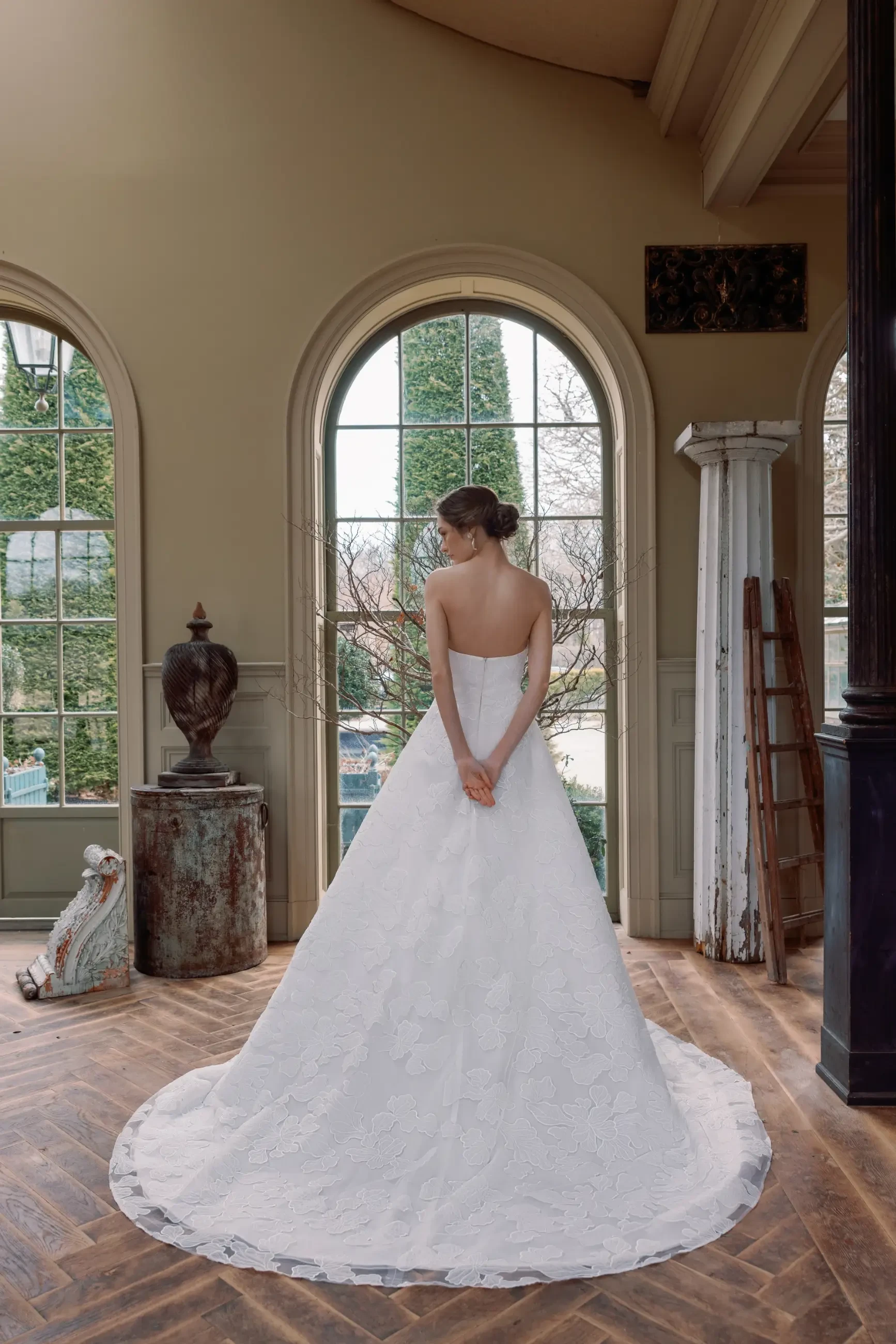 Model is wearing a gown made with delicate Italian embroidered appliqué, showcasing a cat-eye neckline, flowing A-line skirt, and an elegant cathedral-length train.