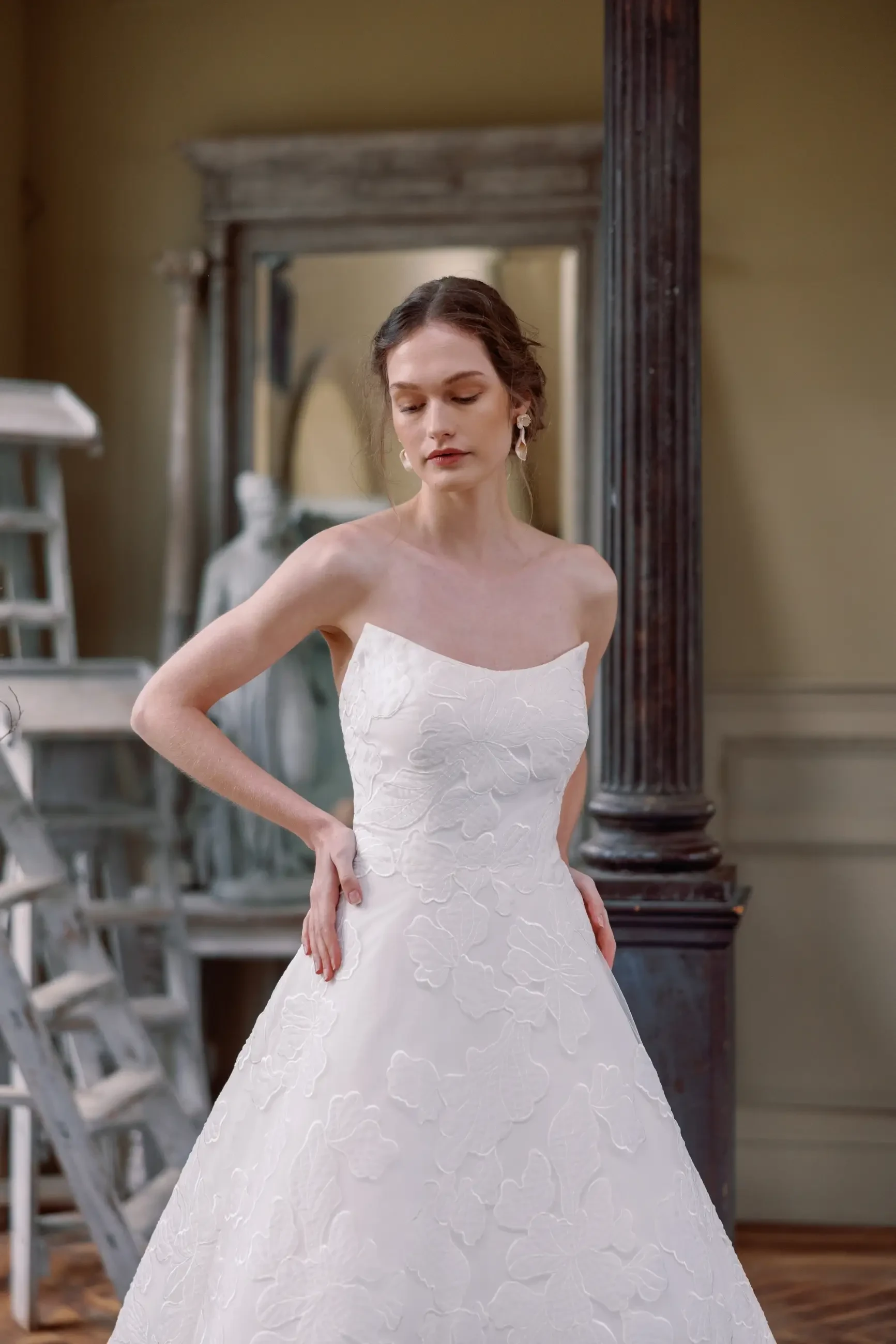 Model is wearing a gown made with delicate Italian embroidered appliqué, showcasing a cat-eye neckline, flowing A-line skirt, and an elegant cathedral-length train.