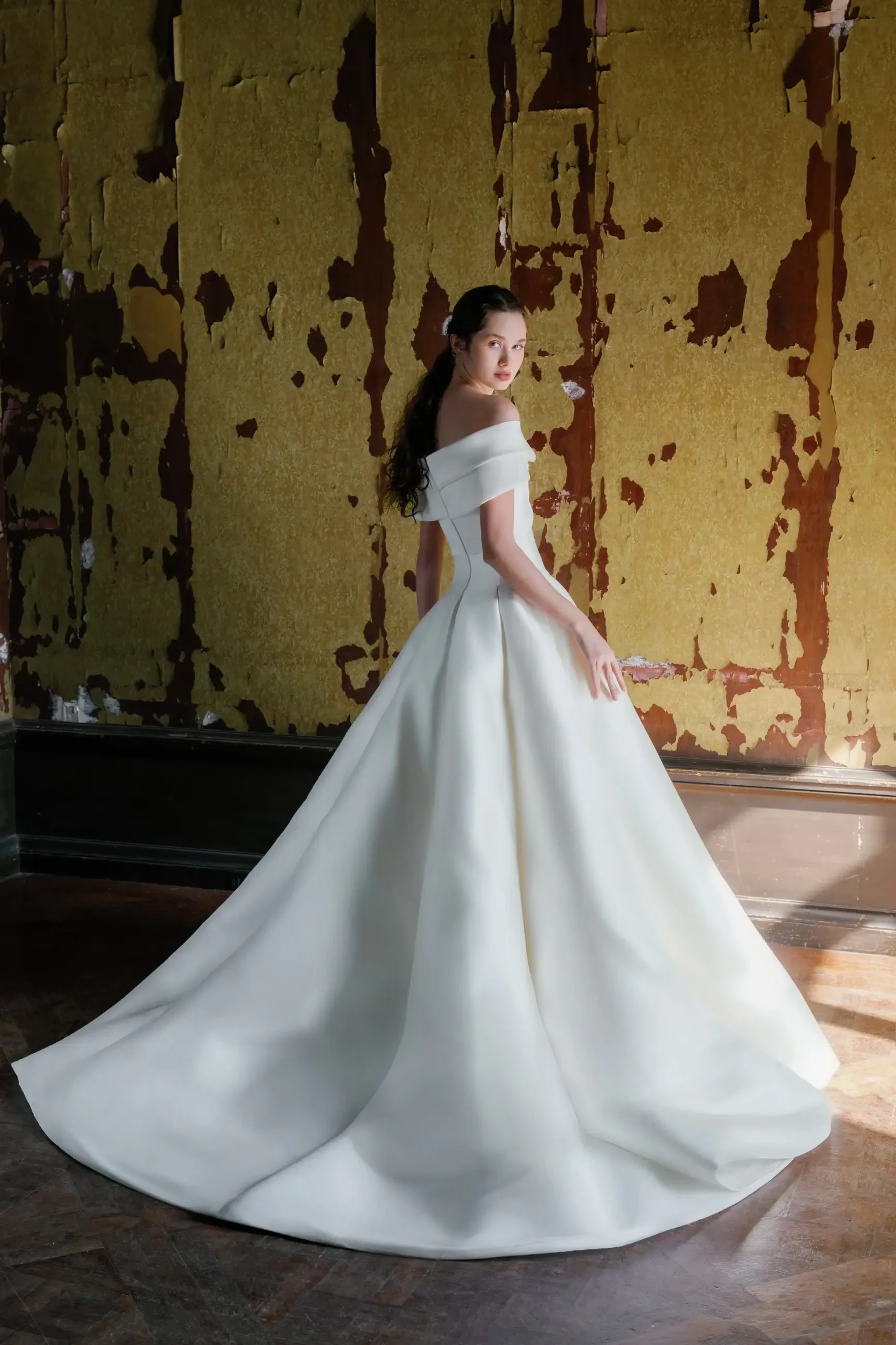 A woman in a white off-shoulder gown stands with her back to the camera, showcasing a long train, against a backdrop of peeling yellow wallpaper.