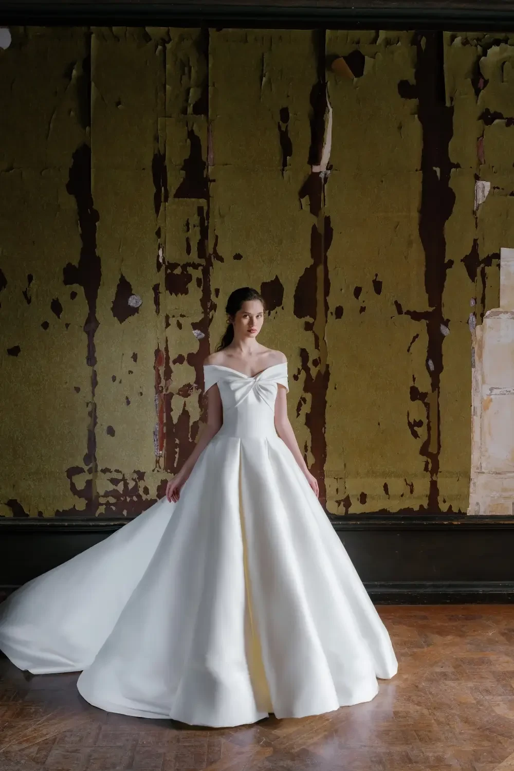A model wearing a white gown with an off-shoulder design and a flowing train, standing in a room with a weathered green wall and wooden floor.