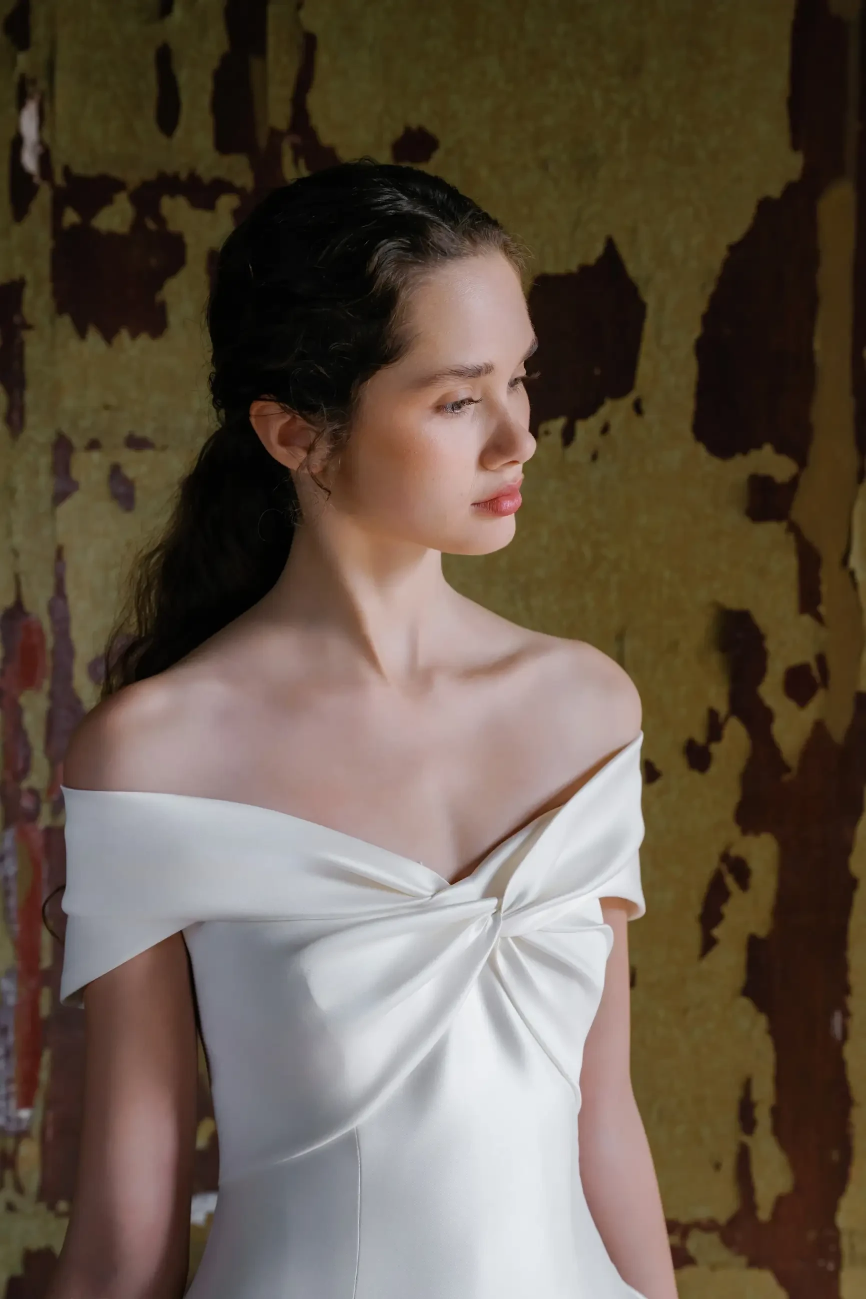 A woman is wearing an elegant off-the-shoulder white gown while standing in a room with peeling yellow paint on the walls.