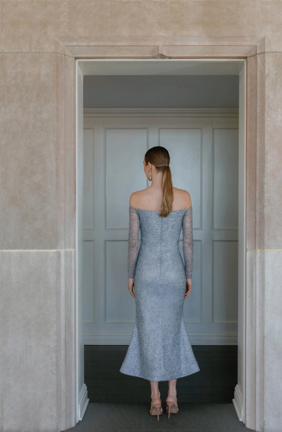 A model wearing a light blue, off-shoulder dress with long sleeves, standing against a wall.