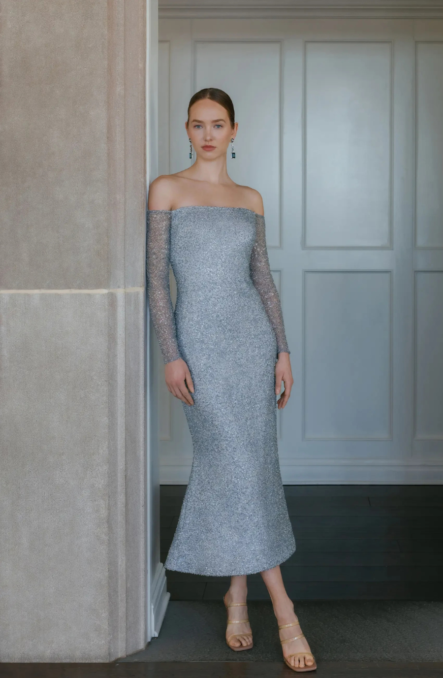A model wearing a light blue, off-shoulder dress with long sleeves, standing against a wall.
