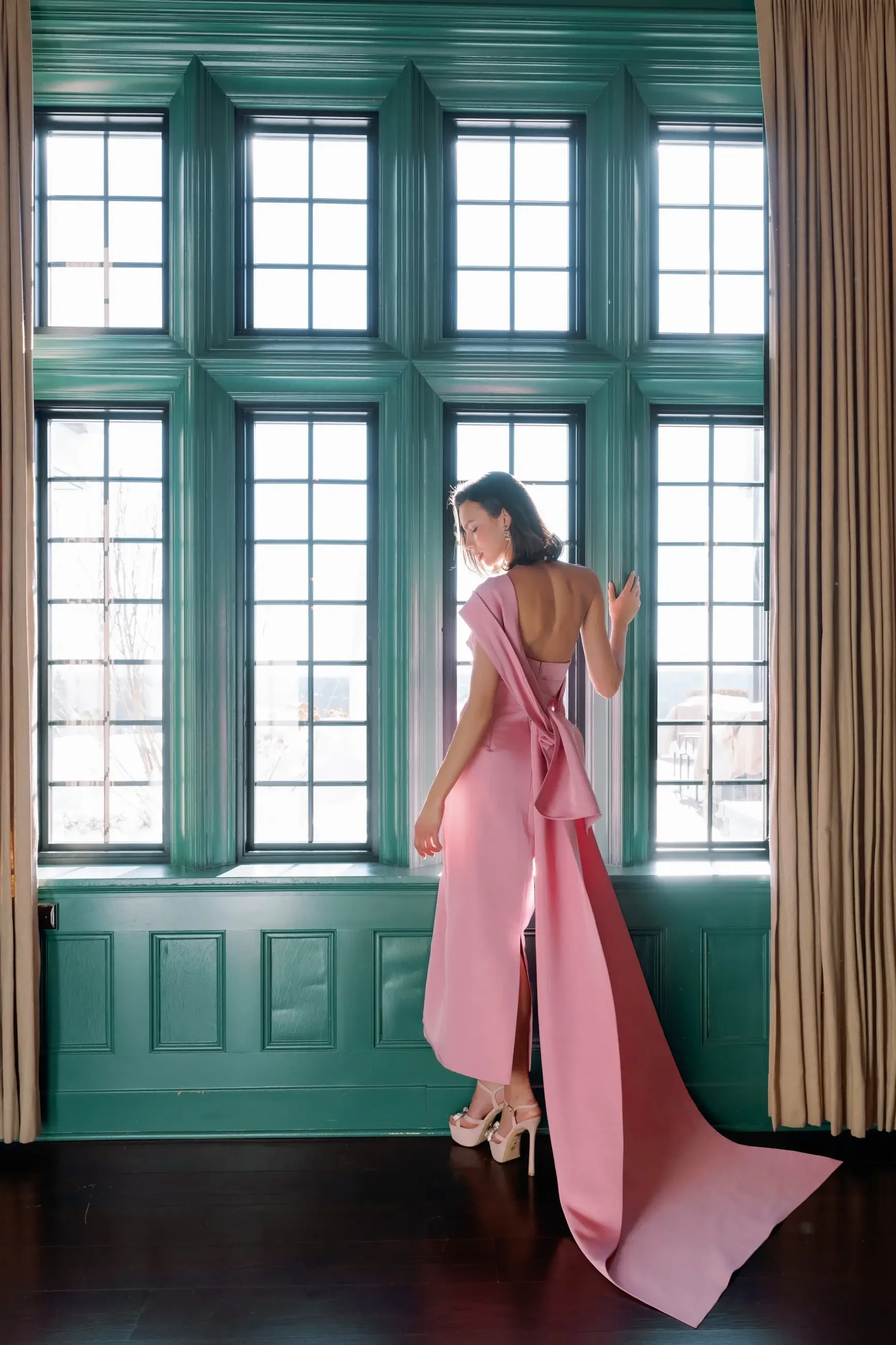 A woman in a pink dress with a flowing train stands by large windows, showing off the dress's back design and silhouette.