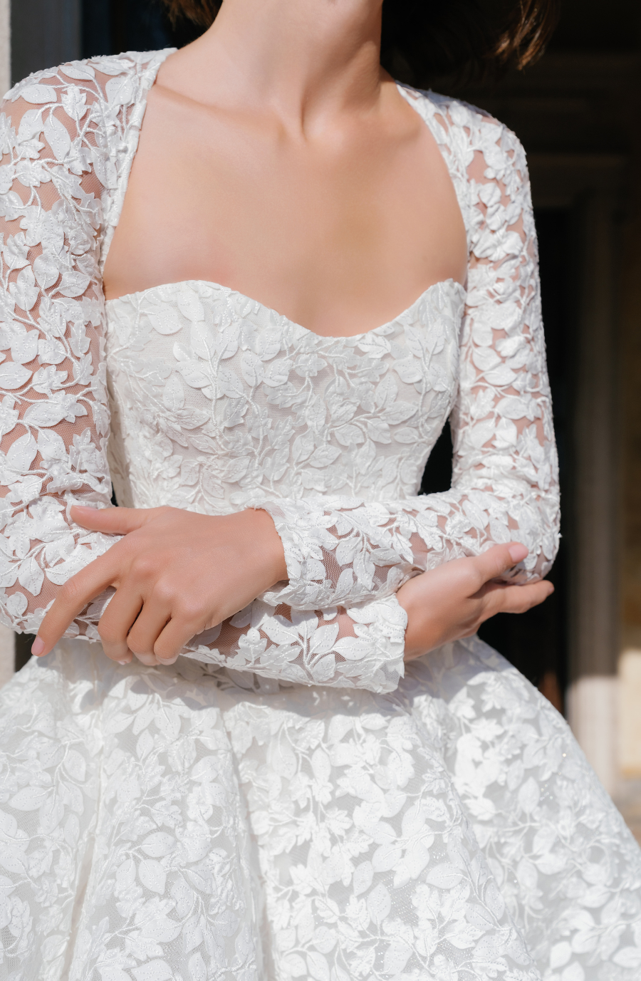 A close-up of a person wearing a white dress with floral lace details and long sleeves, showing only the upper part of their body.