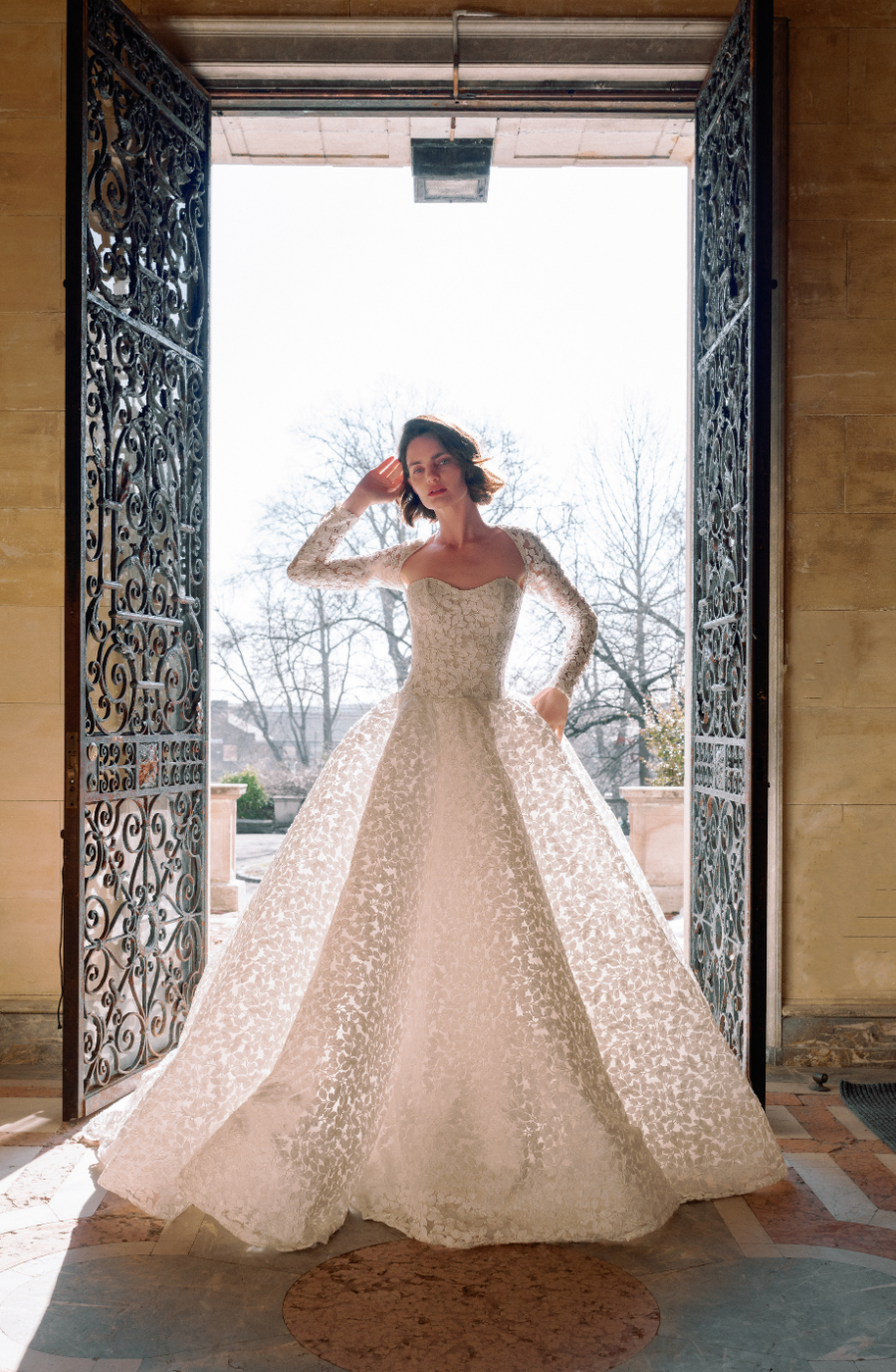 A bride stands in an ornate doorway wearing a white lace wedding dress, with bright sunlight illuminating the scene.